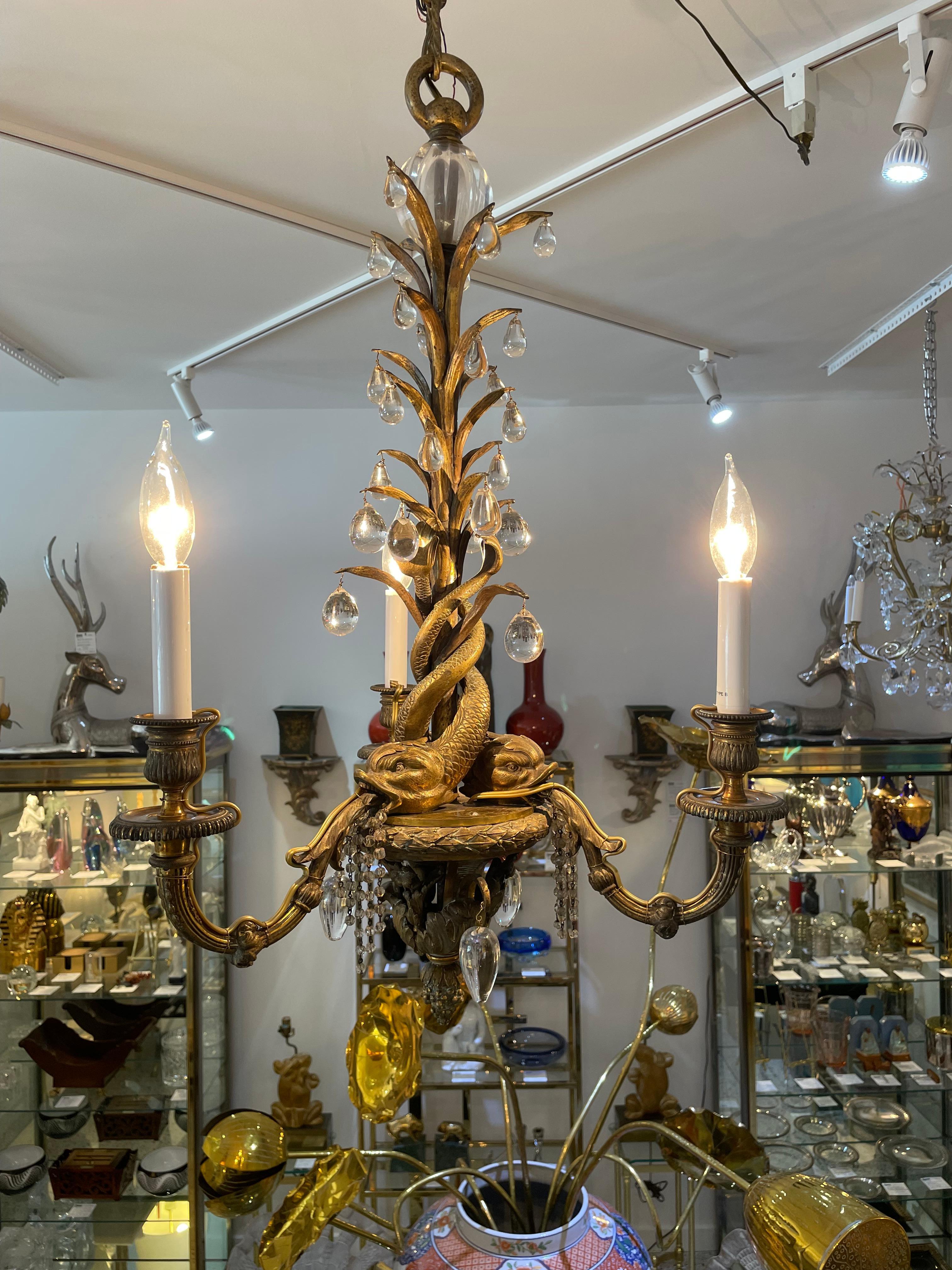 This stylish and chic bronze and crystal chandelier dates to the eary part of the 20th century, and is in the style of Maison Jansen. The piece is detailed with three intertwining sea serpents that have crystal beads spounting from their jaws, and