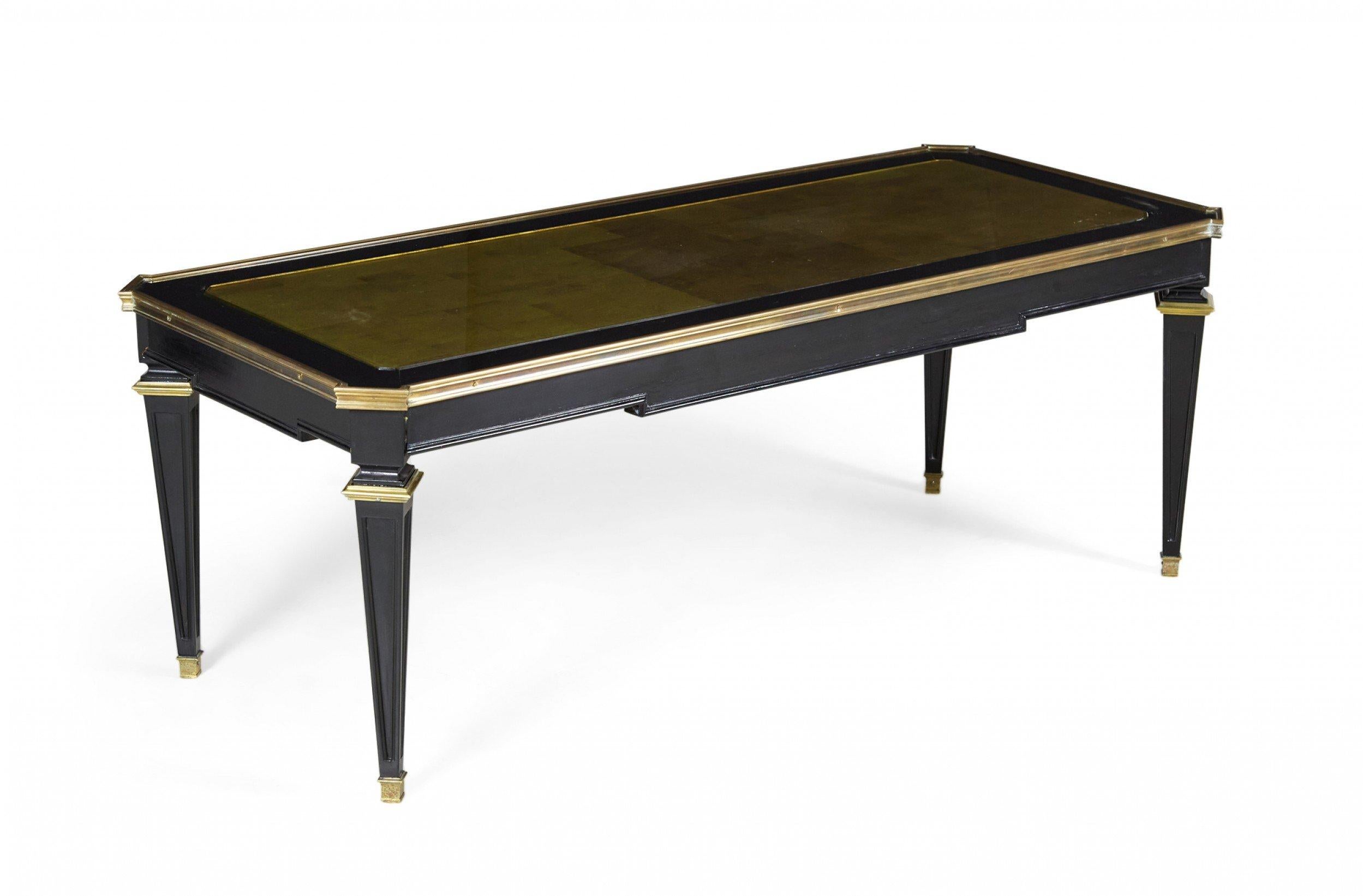 French Mid-Century (1940s) ebonized wooden coffee table with gilt painted trim, an inset gold glass top with geometric pattern, and four tapered square legs ending in brass sabots. (MAISON JANSEN).
 