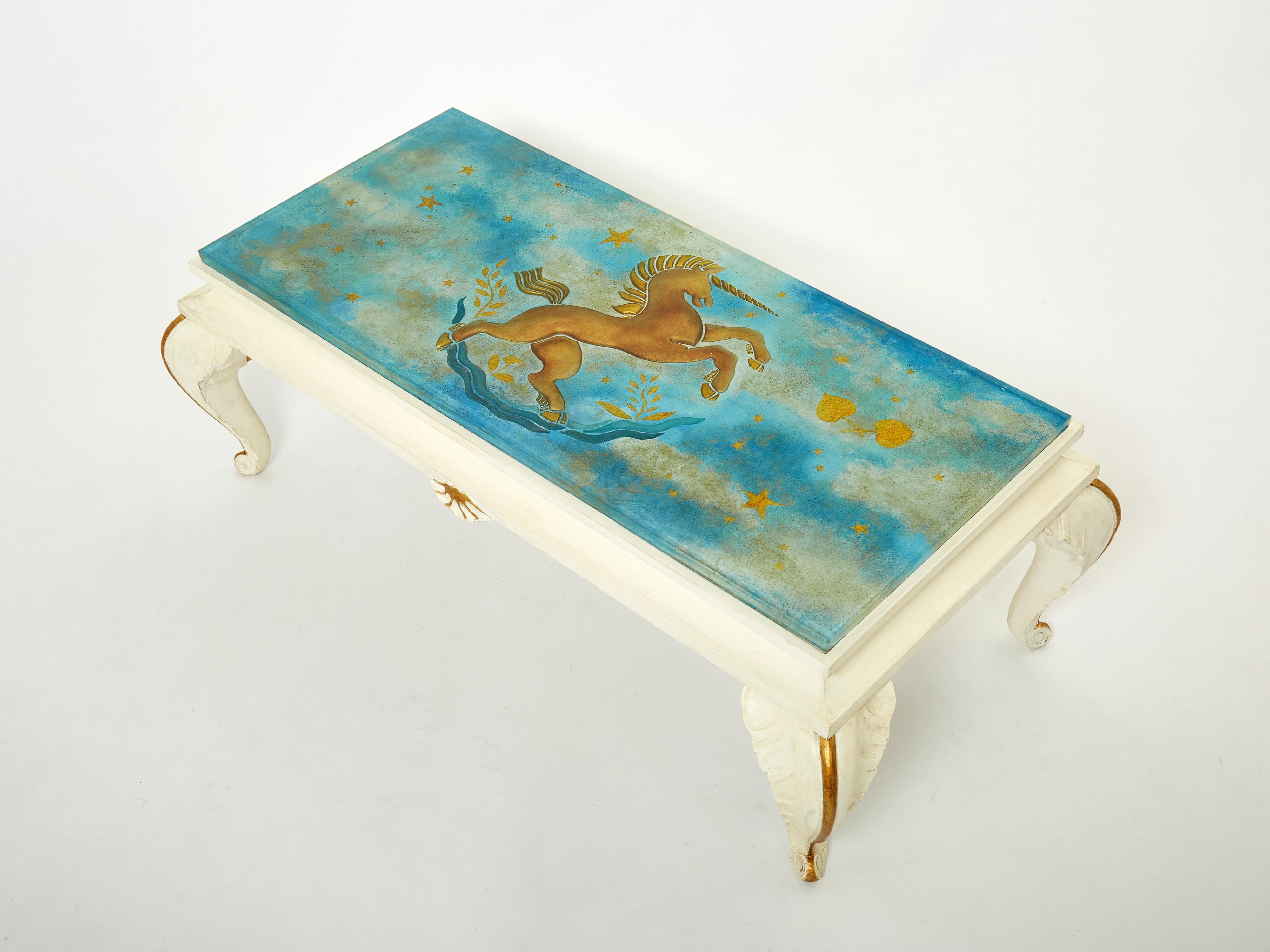 This coffee table by Maison Jansen was made in the early 1950s, with beautifully carved wood feet and structure painted in off white with gilded accents, and an amazing painted glass top picturing a unicorn into a blue clouded sky. The thick Saint