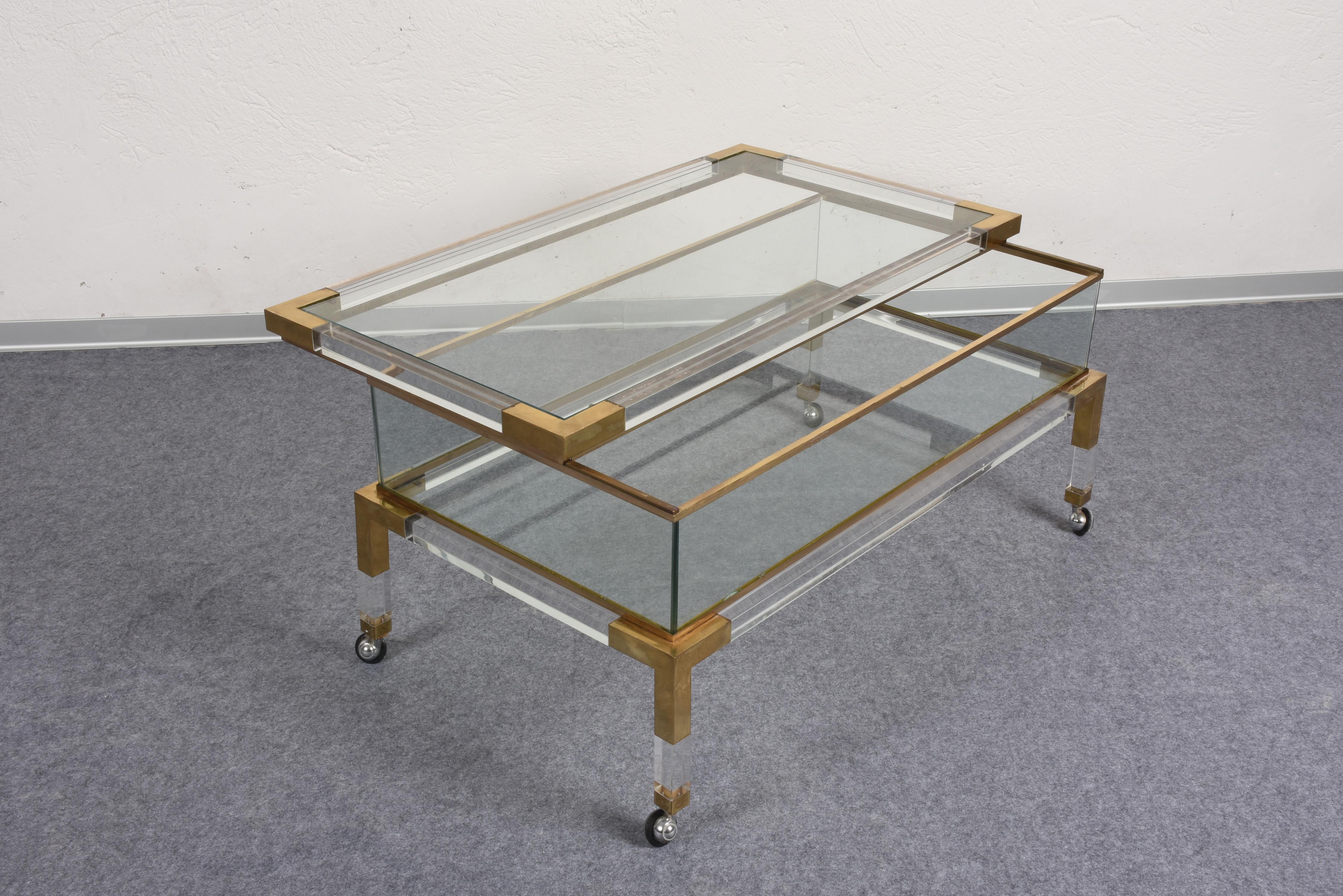 Beautiful midcentury brass and lucite coffee table with sliding shelf and four removable wheels. This fantastic piece was designed by Maison Jansen in France during the 1970s. 

This amazing item is in good condition and has a two-glass top and