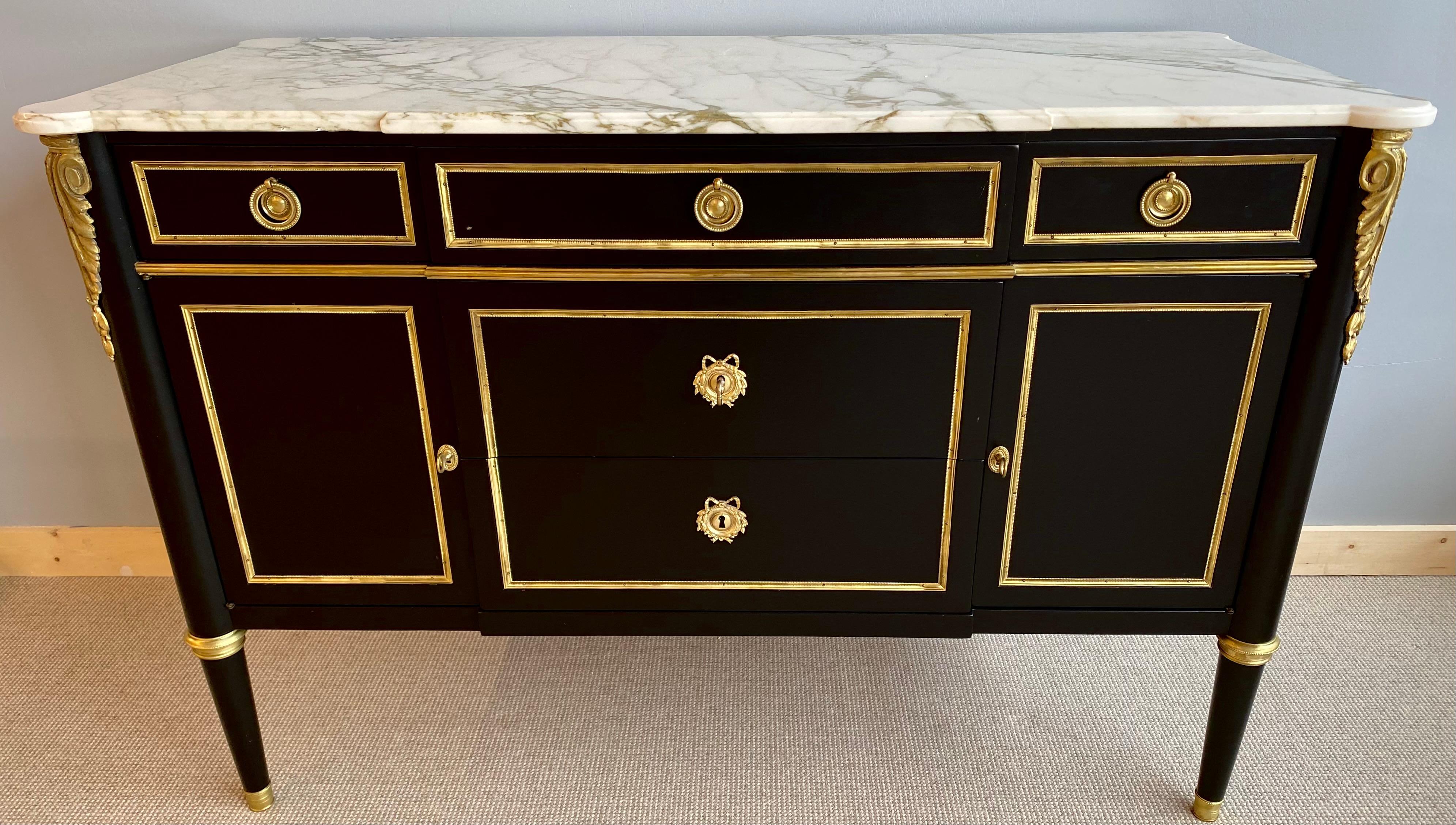 A Hollywood Regency Style Louis XVI fashioned commode by Maison Jansen. The white and gray black veined marble top supported by a ebony case of three drawers with bronze framing over two center bronze framed drawers flanked by two side bronze framed