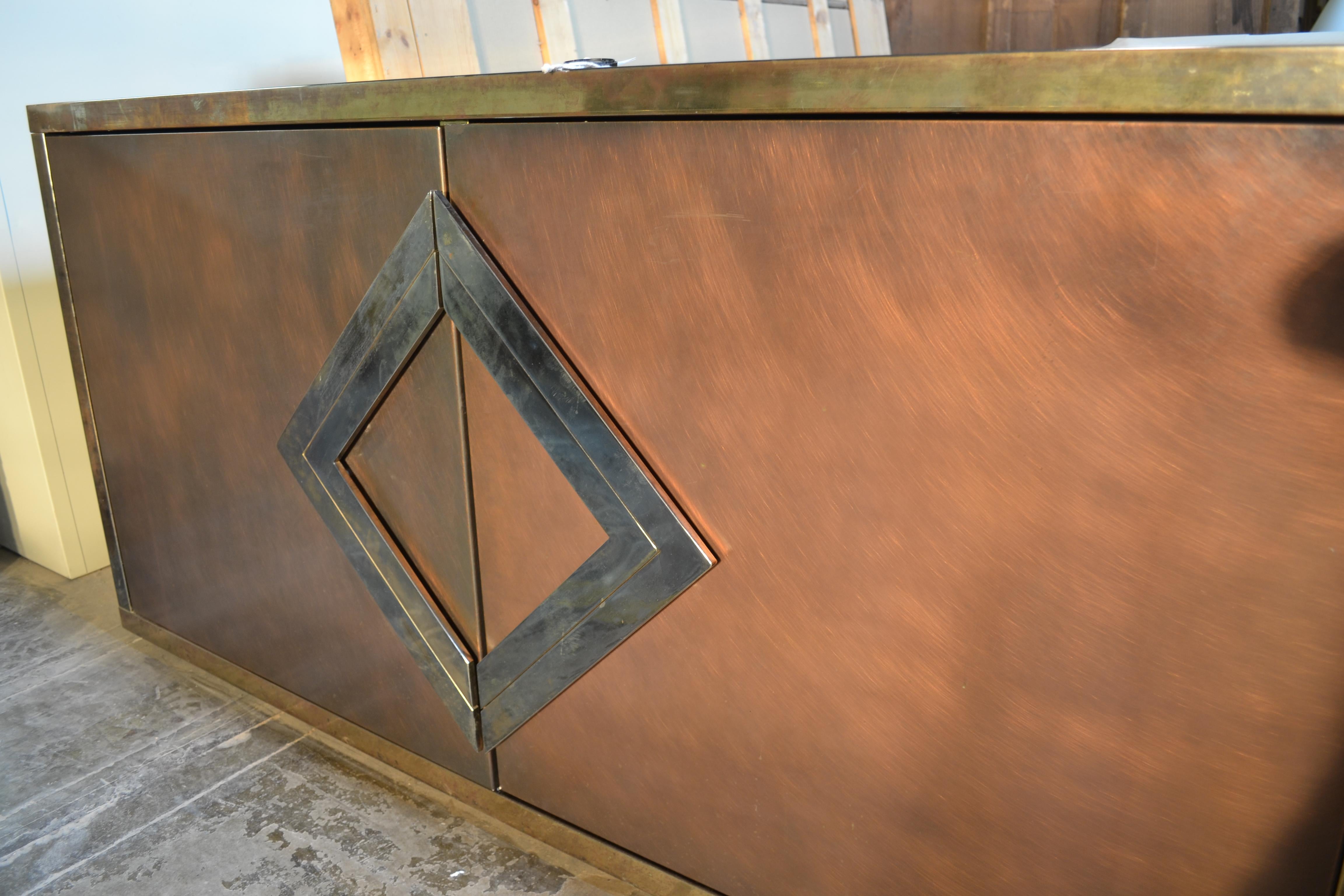 This piece has a superbly worn finish. It has a body of textured copper banded in chrome and highlighted with boldly geometric door pulls. There are two cabinet areas, each with a glass shelf. It's in good condition with some scuffing to the metal
