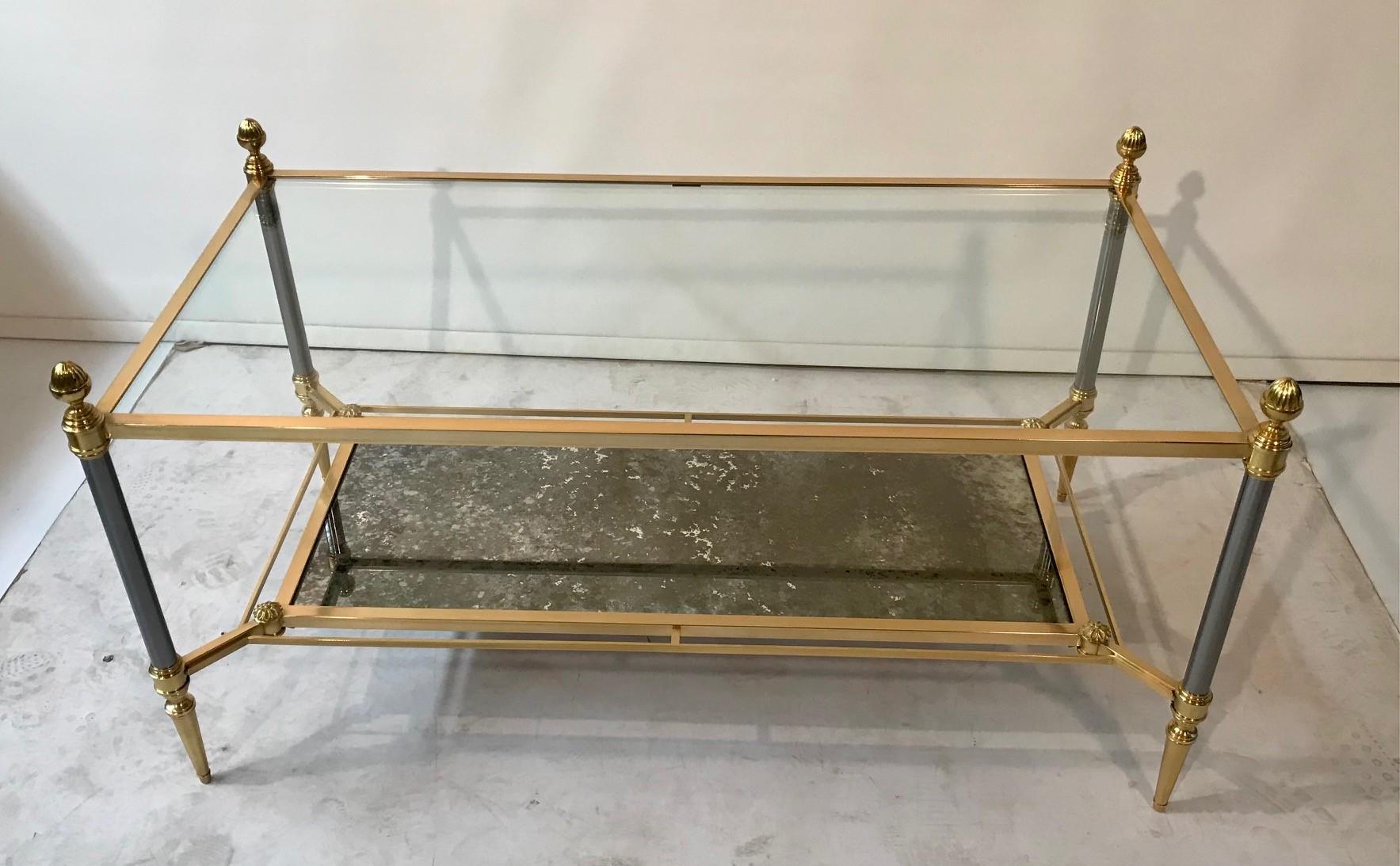 This two tier coffee table is supported by four brass and burnished steel uprights, each with a lobed acorn finial, The upper tier is clear plate glass, the lower (and smaller) is finished with a patinated antique finished mirror, each is framed in