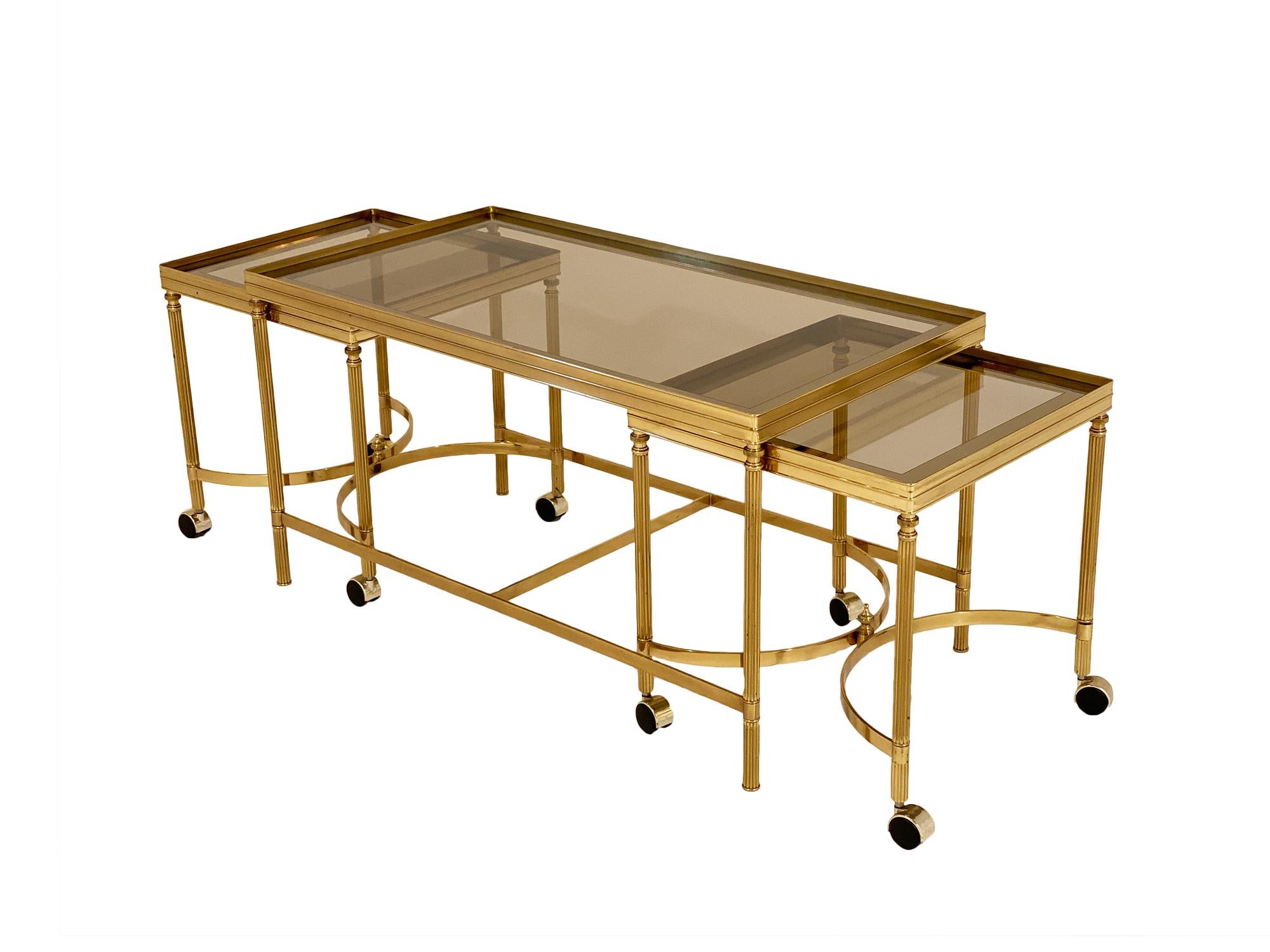 Maison Jansen nesting coffee table set from France. Made of brass with lightly smoked glass; this set by the iconic Parisian company Maison Jansen has classic details and beautiful quality. The main table has an H stretcher and two side tables on