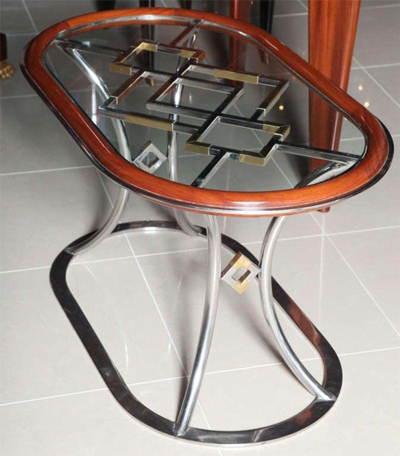 The mahogany banding with chromed steel and brass plated steel geometric cut-out motifs under a glass top, on curved legs and an oval stretcher, designed by Alain Delon for Maison Jansen in 1974.