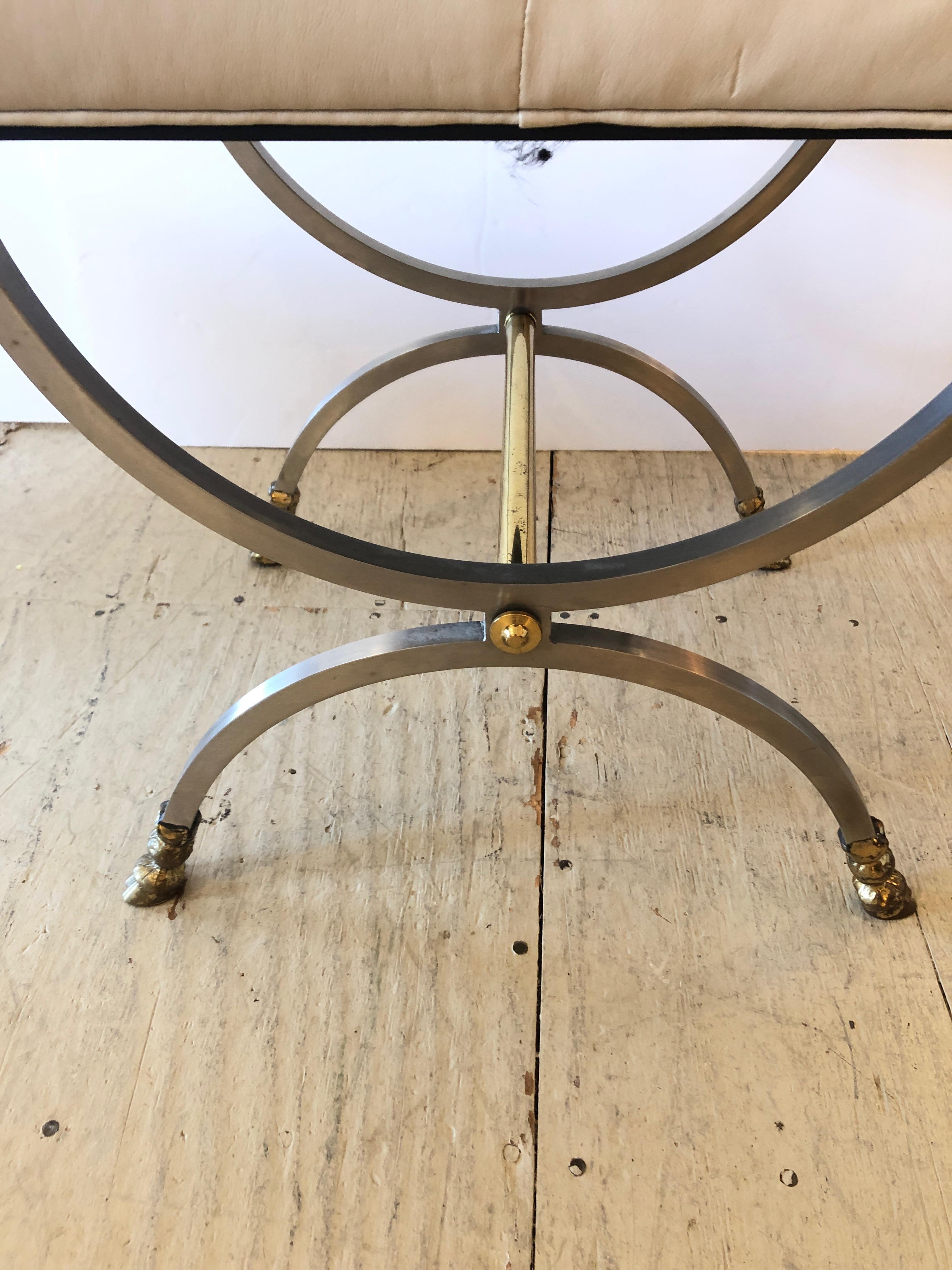 A sexy Mid-Century Modern ottoman or bench having steel and brass double U-shaped legs on each end with stretcher and Maison Jansen esque hoof feet. The attached cushion is supple creamy bone leather.