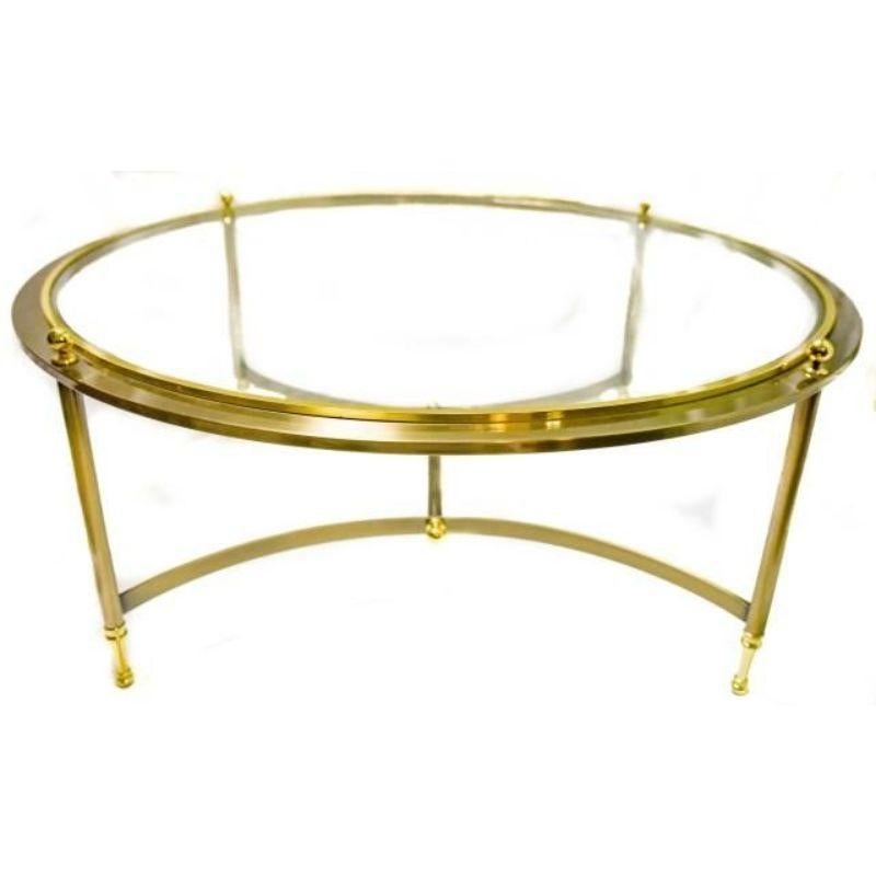 A vintage Maison Jansen style round glass top coffee table with brass and gold tone frame. The round table has four straight legs, each topped with a traditional finial at the table top. The table has curved stretchers meeting in a single I shape at