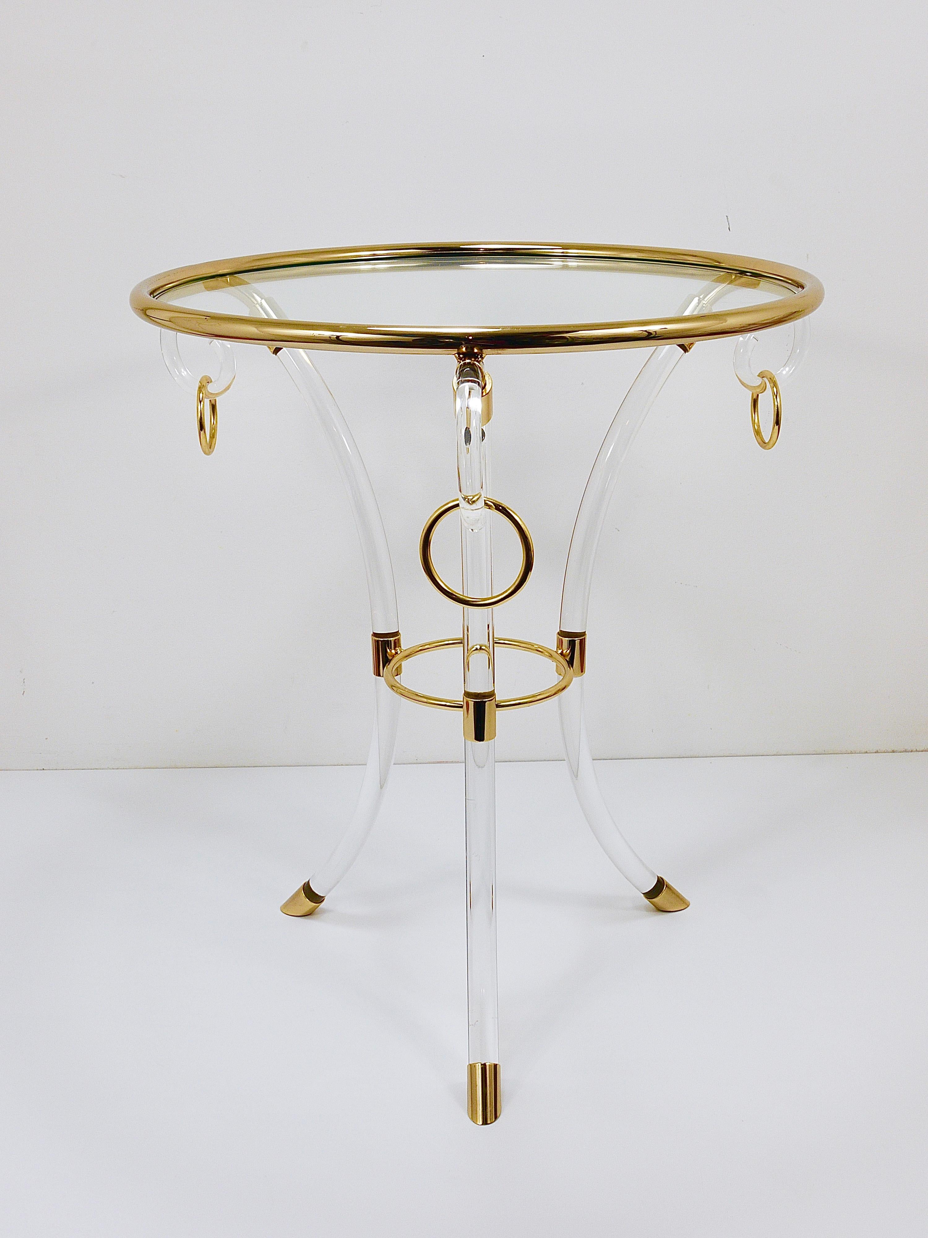 This French round coffee table / side table from the 1970s captures the essence of the Hollywood Regency era. Crafted from clear acrylic Perspex glass / plexiglass and gilded metal, it features elegant golden details.  The three curved lucite legs