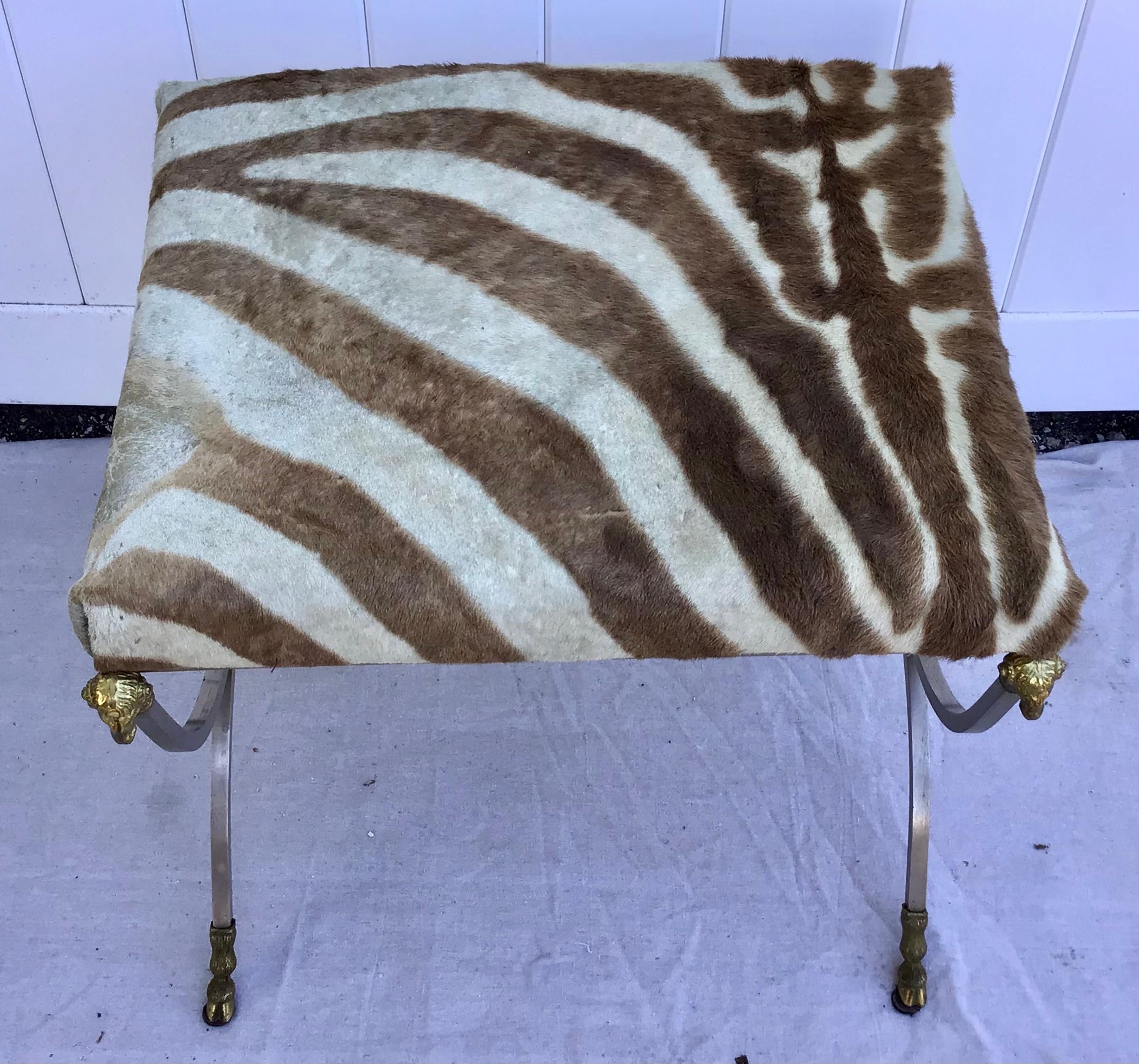Stunning lines on this Maison Jansen of Paul style bench. The zebra hide upholstered seat over iron form stretcher with brass Ram's head and hoof feet.