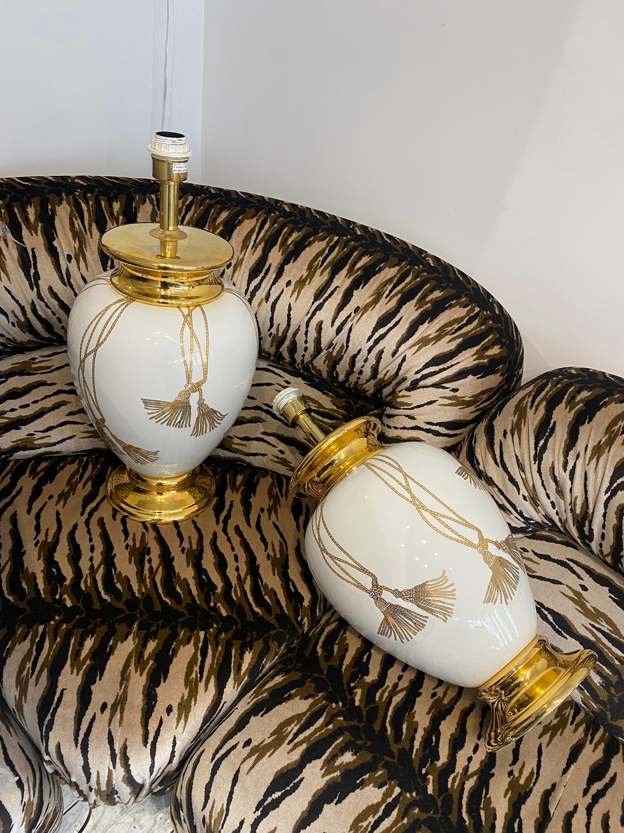 An impressive signed pair of 1990s table lamps by Maison Le Dauphin in white ceramic body and 24k gold plated base and neck. 
The body is decorated with gold strings of tassels giving the illusion that are tied on the neck of the lamp. An elegant