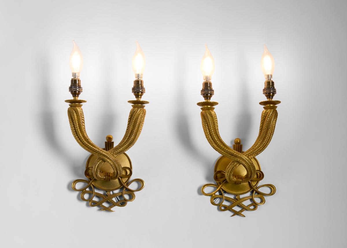 A charming pair of gilt-metal sconces underlain with delicate crisscrossing design reminiscent of script and possessing two symmetrical arms which extend from the fixtures' centers like smoke twisting gently from a flame. Dating from the middle of