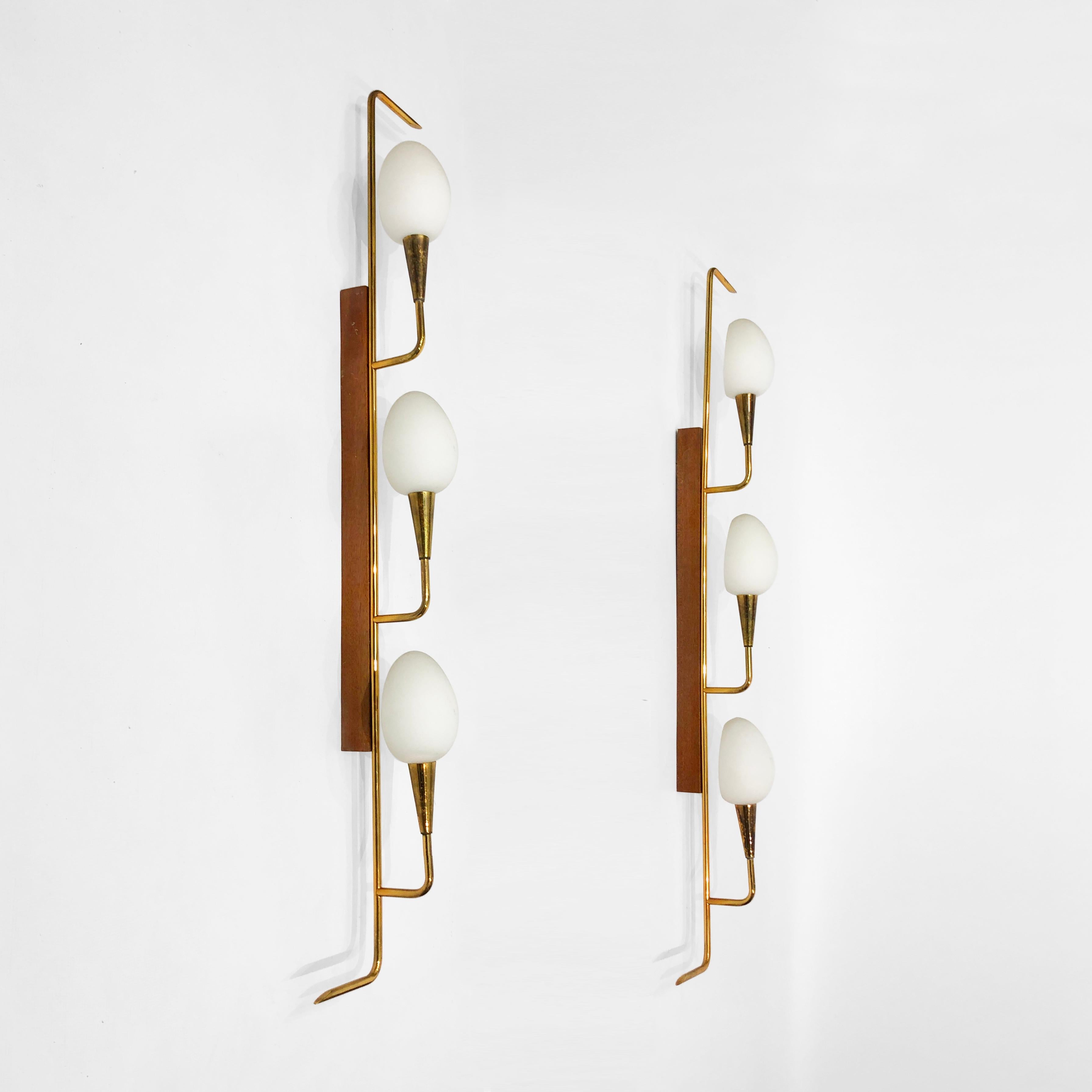 An impressive pair of 1950s mid-century wall lights by Maison Lunel, imported from an estate in Bari, Italy. A teak spine supports a brass frame, from which protrudes three branch-like brass cones, each holding an egg-shaped glass shade. The brass