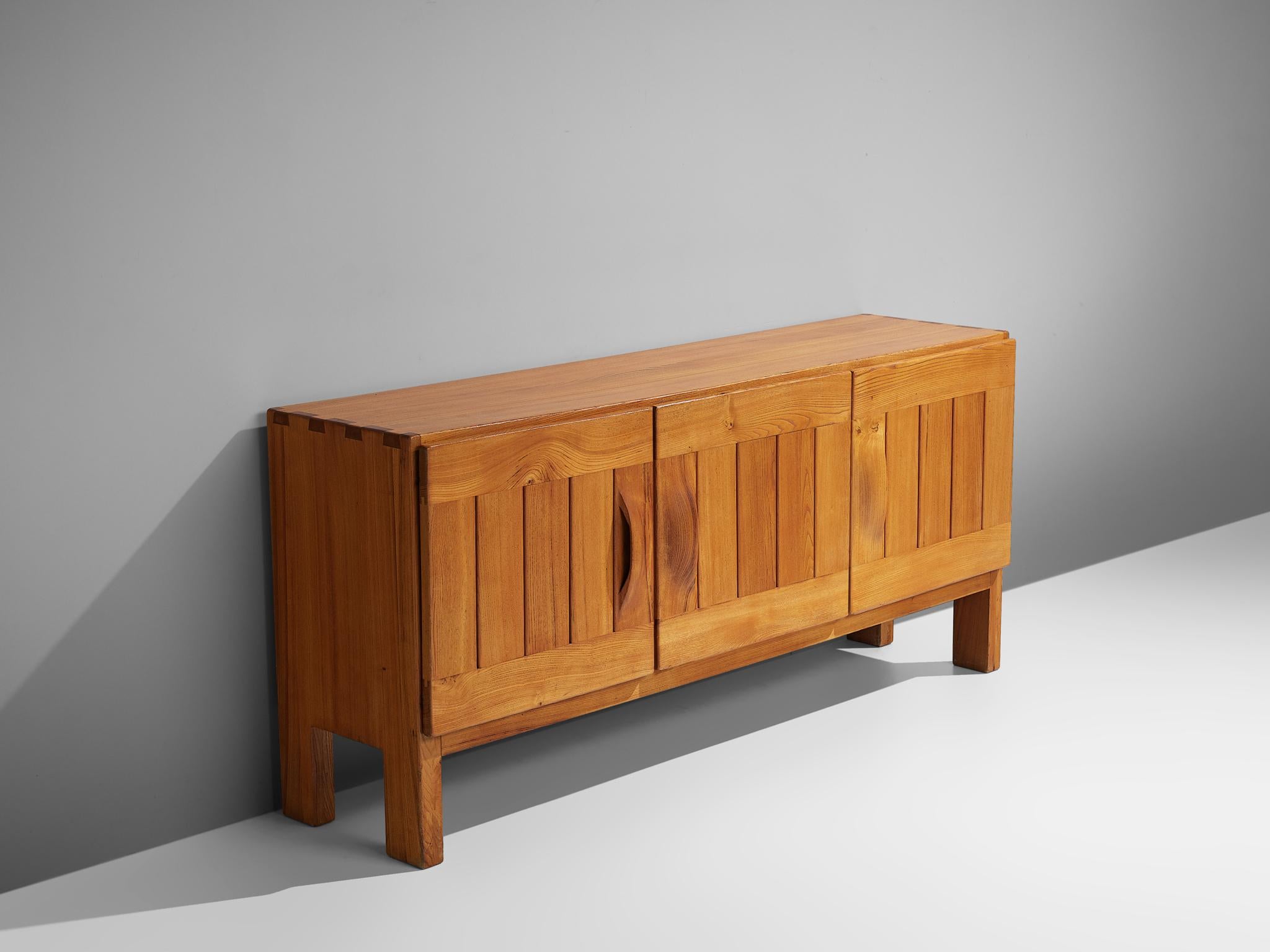 Maison Regain, sideboard, elm, France, 1960s.

This elm credenza combines a simplified yet complex design combined with nifty, solid construction details that characterize Maison Regain's designs. The three well balanced, sturdy doors are geometric