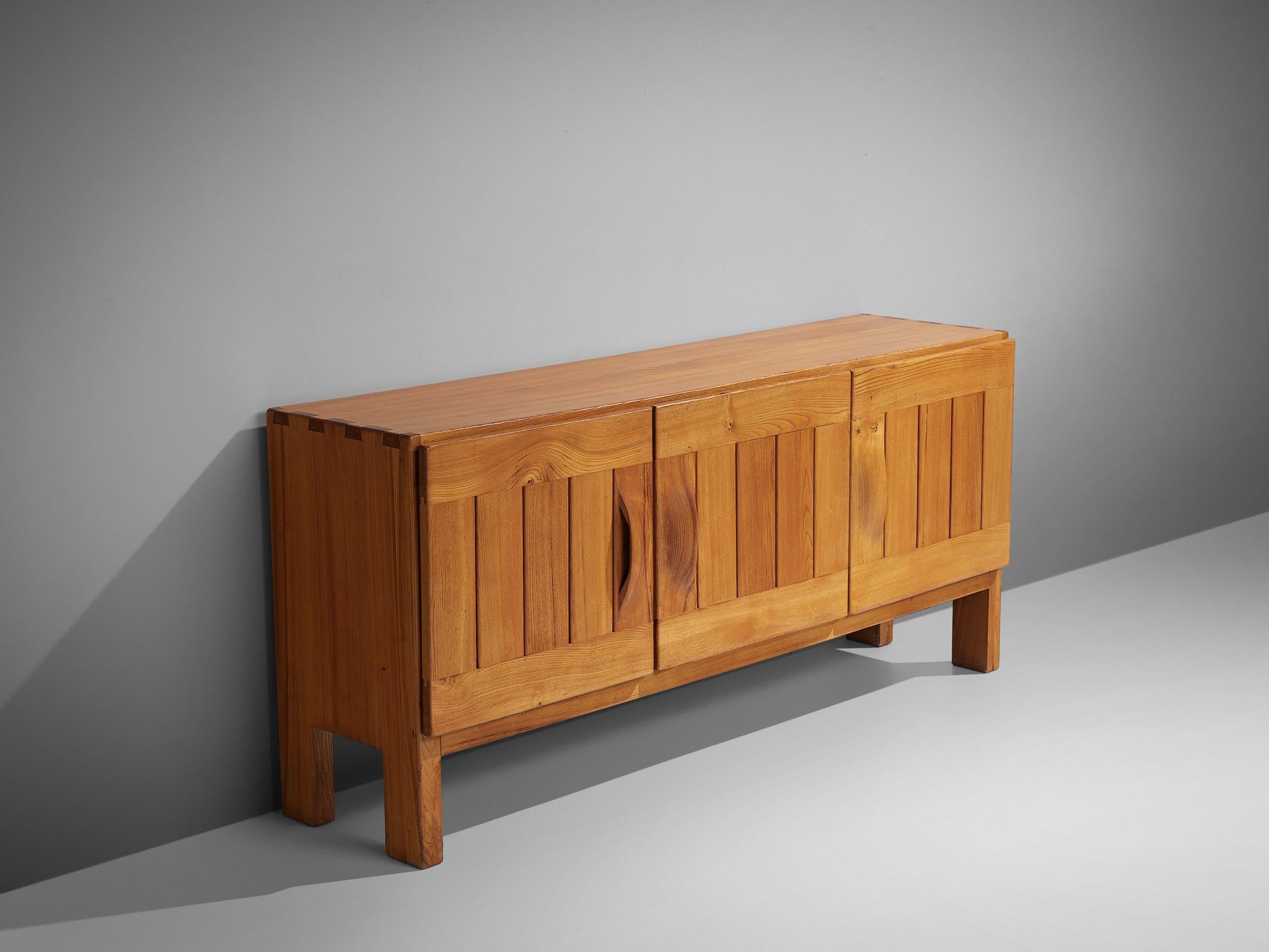 Maison Regain, sideboard, elm, France, 1960s.

This elm credenza combines a simplified yet complex design combined with nifty, solid construction details that characterize Maison Regain's designs. The three well balanced, sturdy doors are