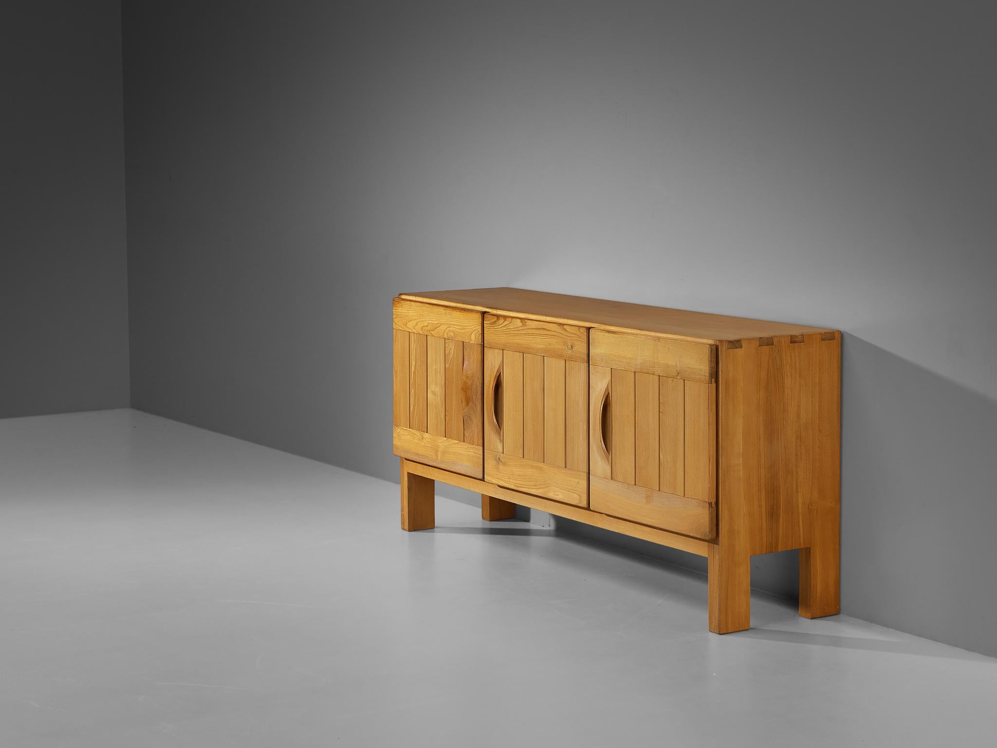 Maison Regain, sideboard, elm, France, 1960s.

This sideboard by Maison Regain combines a simplified yet complex design combined with nifty, solid construction details that characterizes its design. The three well-balanced, sturdy doors are