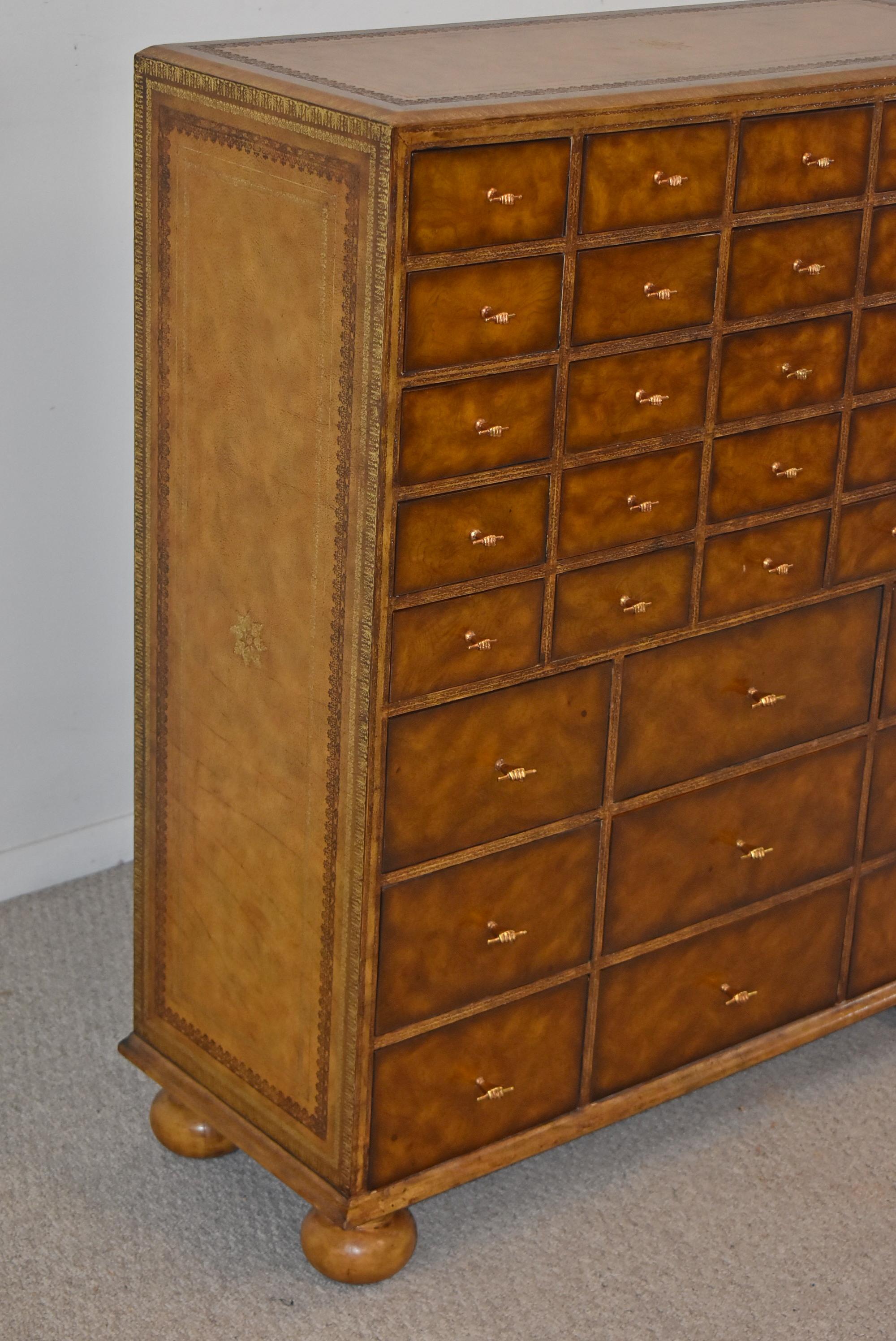 This beautiful Maitland Smith apothecary cabinet with 34 drawers is wrapped in tooled leather. The drawers fronts are made from burled walnut and are adorned with copper tone pulls in the shape of a hand. This piece is in excellent pre-owned