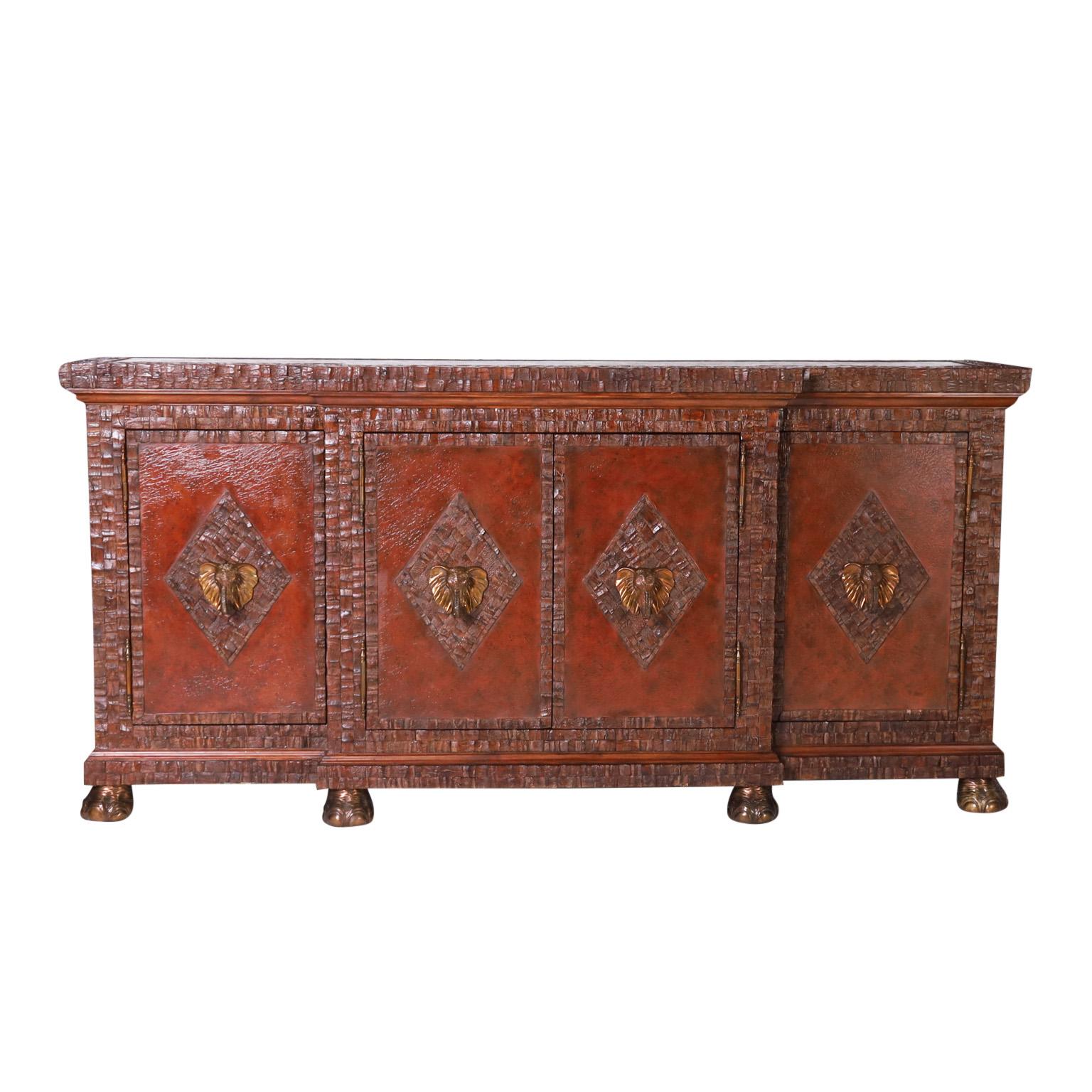 Impressive midcentury Maitland-Smith sideboard or wine cabinet with a textured bronze top in a coconut border on a four door case having bronze elephant head handles in coconut panels on leather backgrounds with coconut shell frames. The mahogany