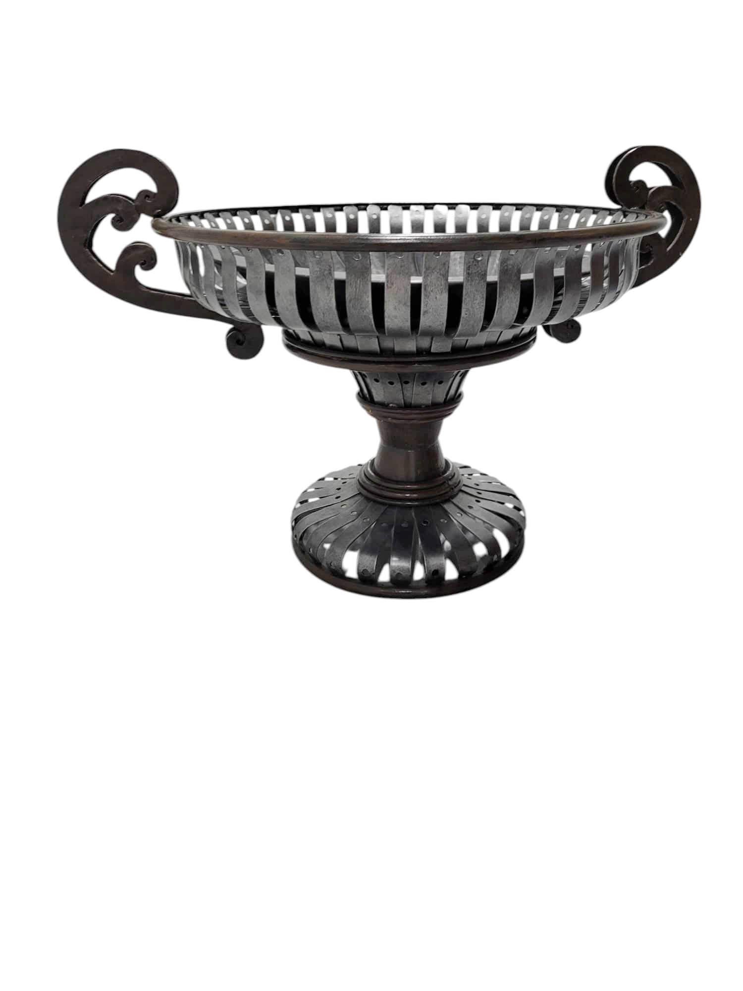 Questo centrotavola Maitland Smith colpisce per le sue dimensioni e la sua presenza, fondendo l'artigianato industriale con l'eleganza neoclassica. Il corpo reticolato, realizzato a mano con strisce di metallo rivettate, abbina i toni del bronzo