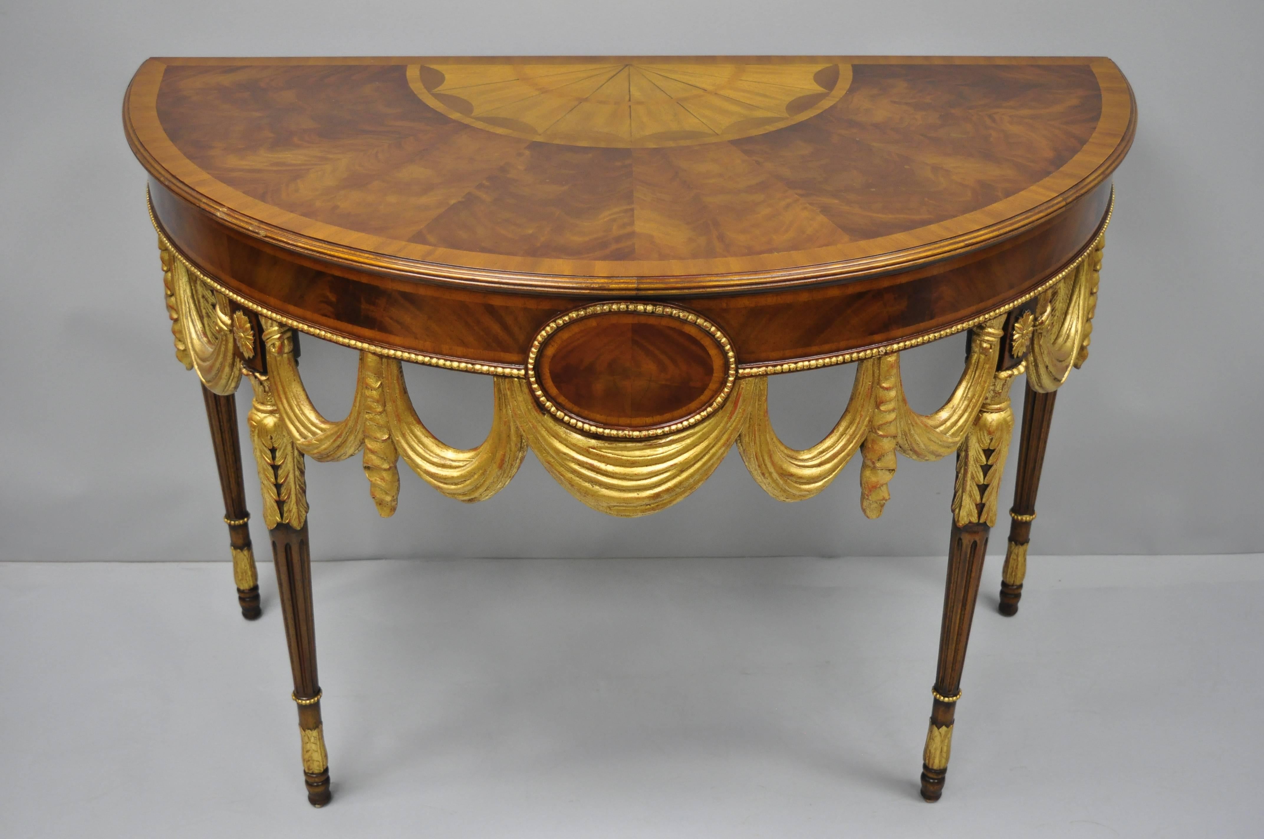 Maitland Smith half round inlaid demilune console table. Item features a crotch mahogany sunburst inlaid top, gold gilt finish, drape carved skirt, beautiful wood grain, original label, tapered legs, great style and form, circa early 21st century.