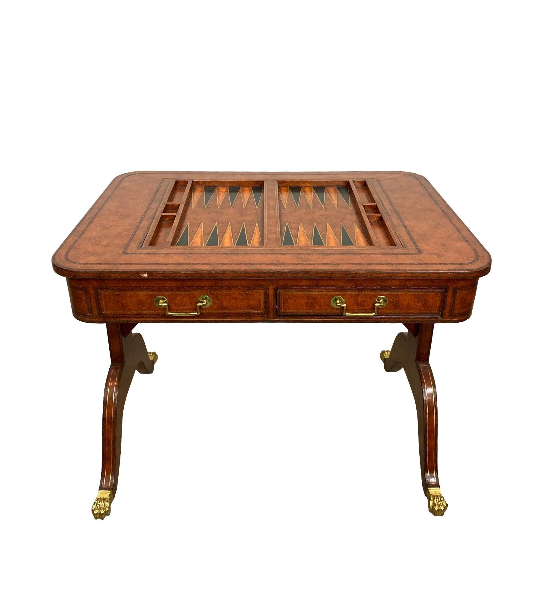 Maitland-Smith Regency-style chess/backgammon board table, covered in tooled and embossed leather, with gilded highlights, the single stretcher bar above Regency-style legs, with gilded leather stringing, on brass castors with hairy paw toe covers,