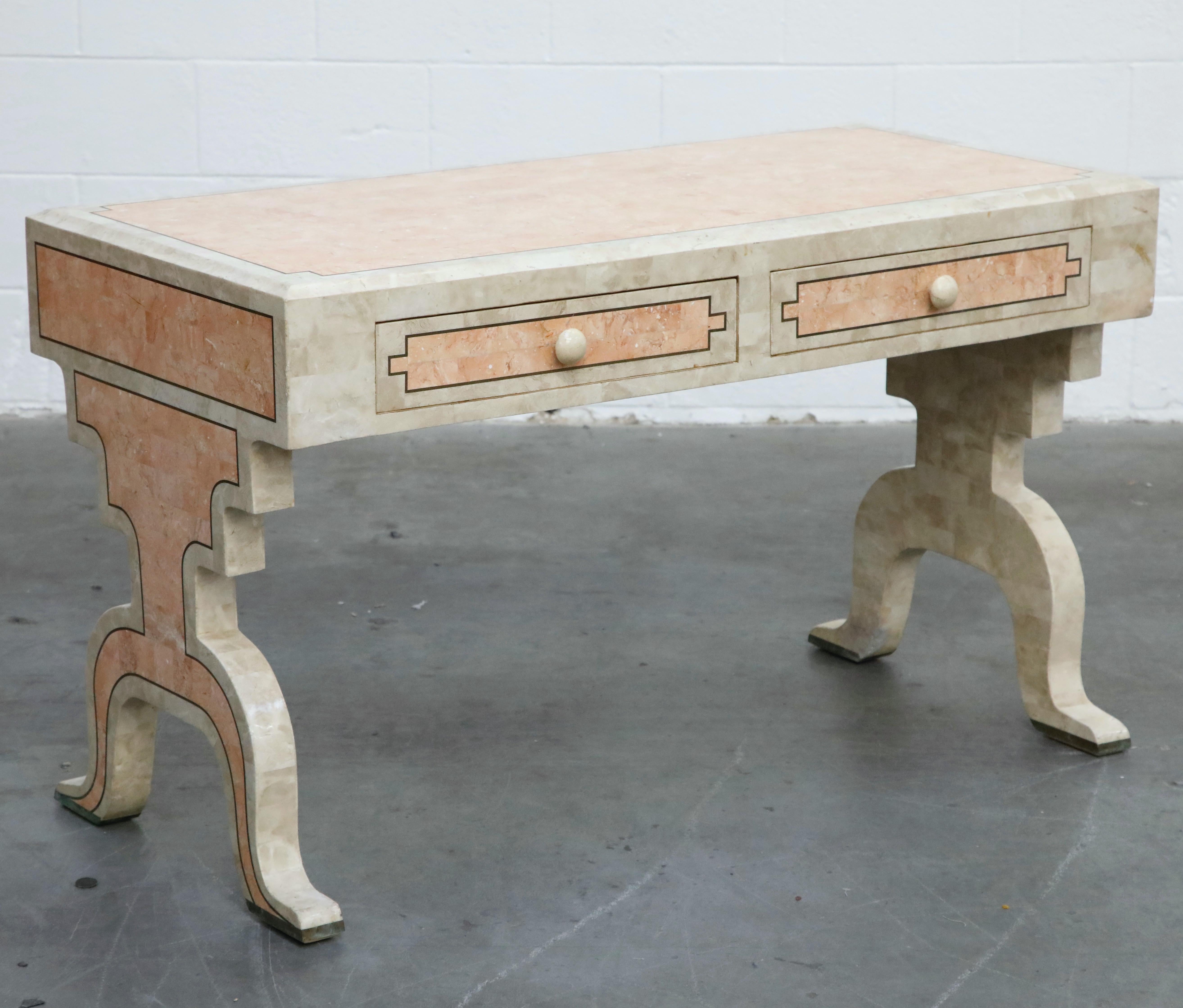This gorgeous Maitland Smith two-drawer desk features impressive tessellated pink marble and stone with brass inlays. This beautiful circa 1980s desk is in an Art Deco and Hollywood Regency stye with shaped legs and compact size ideal for smaller