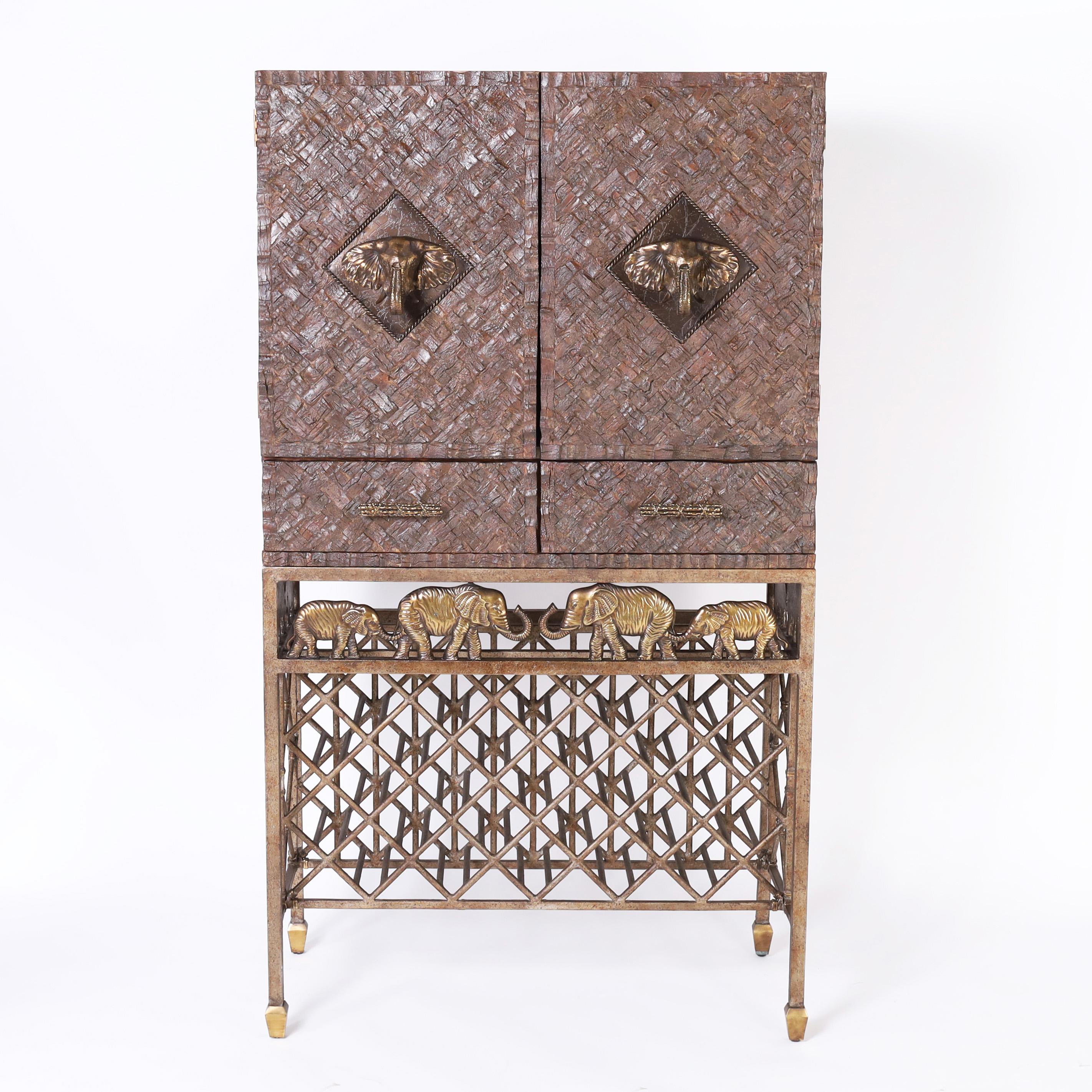 Impressive mid century bar cabinet featuring an upper case crafted in coconut shell with two doors having bronze elephant head handles and an illuminated interior lined in mahogany with a travertine surface over two drawers on an iron base with