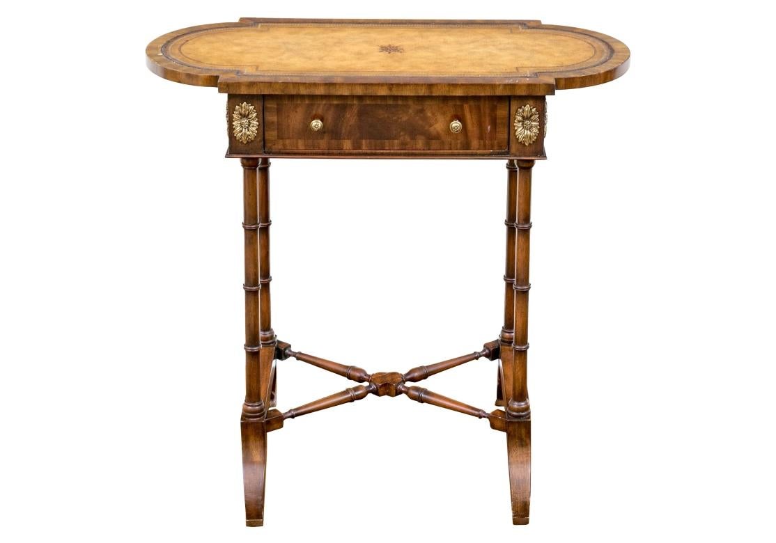 Maitland-Smith accent table featuring a hand-tooled brown leather top with breakfront sides overhanging a single drawer in figured warm-toned medium brown veneers. Detailed with ornate gilt oval floral medallions heralding faux bamboo cylindrical