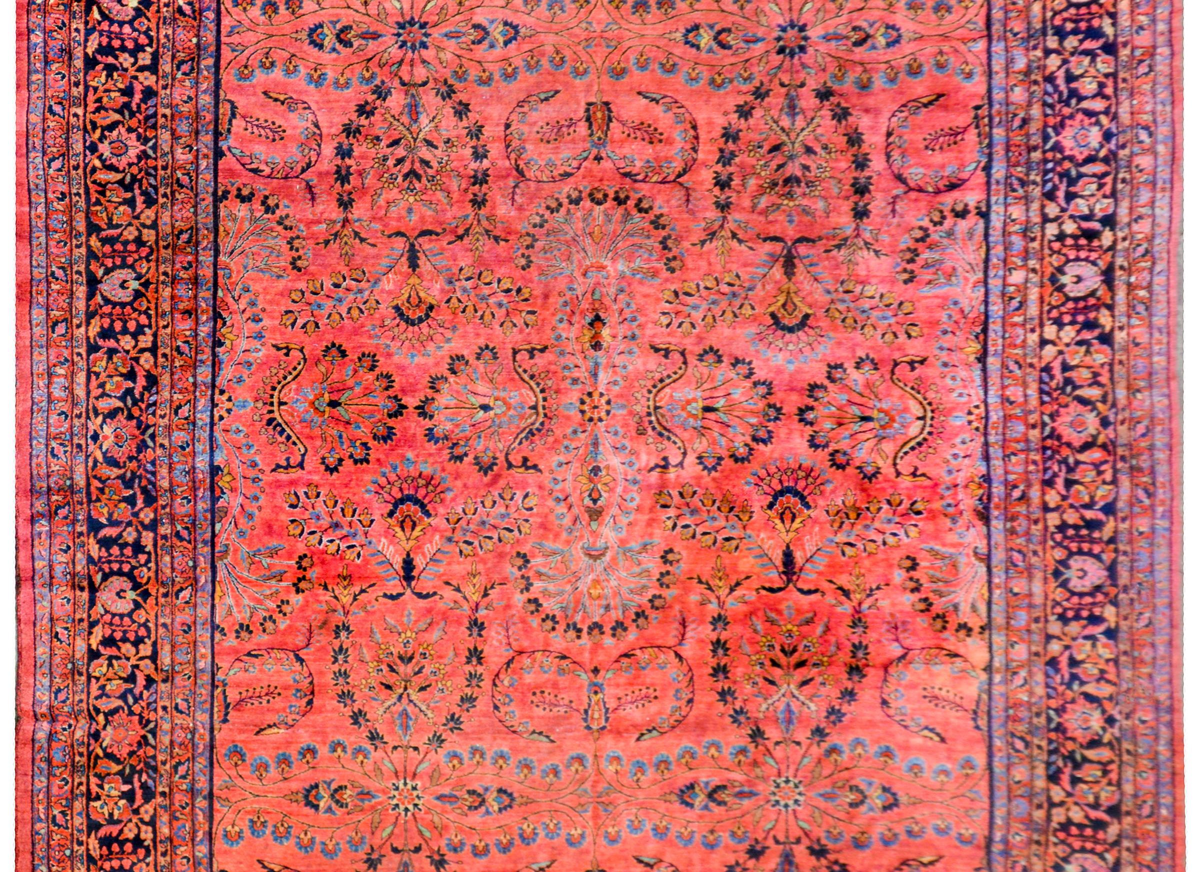 A majestic early 20th century Persian Sarouk Mohajeran rug with a large-scale mirrored floral pattern with myriad flowers and vines woven in light and dark indigo, crimson, and orange, on a coral colored background. The border is beautiful with a