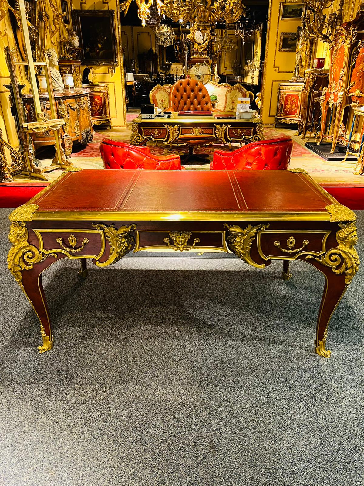 Root veneer on solid beech and oak. Extremely finely chiselled bronze. Slightly protruding, profiled tabletop and gold-imprinted leather insert, flanked by a grotesque mask on each corner. Three-wheeled frame base with wide knee-section on four