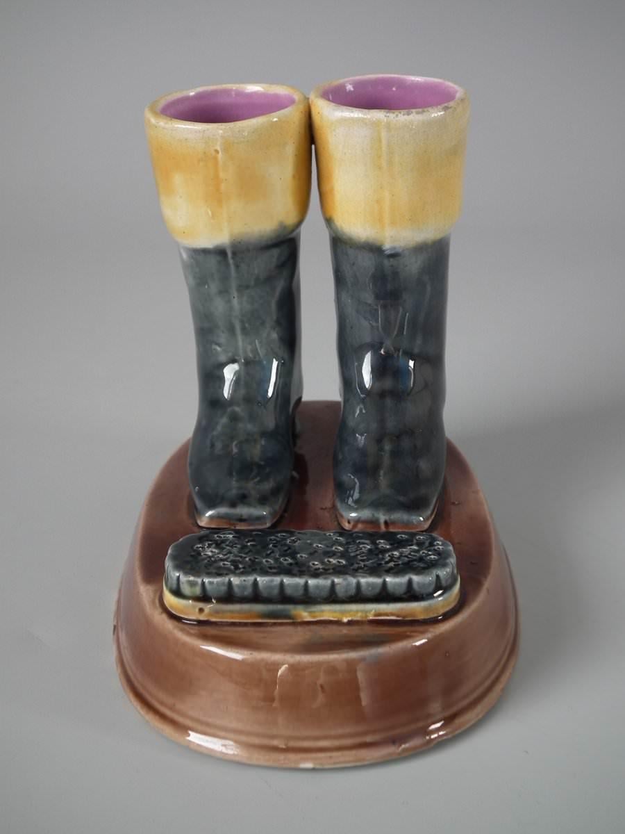 Majolica match striker which features a pair of boots next to a polishing brush on an oval base. Coloration: brown, black, ochre, are predominant. Bears a pattern number, '3'.