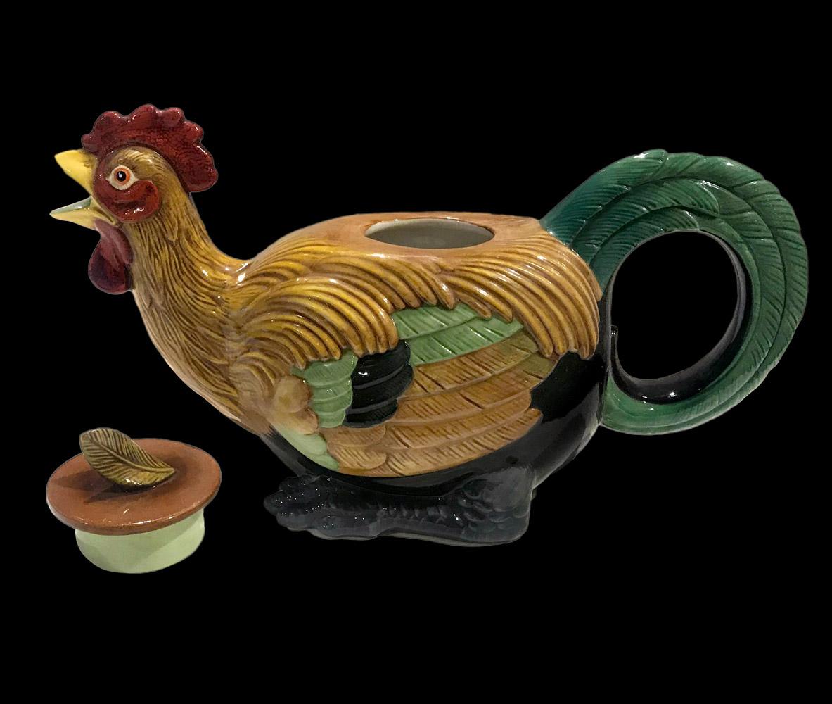 Cockerel Majolica Teapot. He is singing and its beak is open and forms the spout. Its legs are slightly folded beneath him and its feathers are ochre and green, long and the tail ones are forming the handle. A small feather adorns the removable