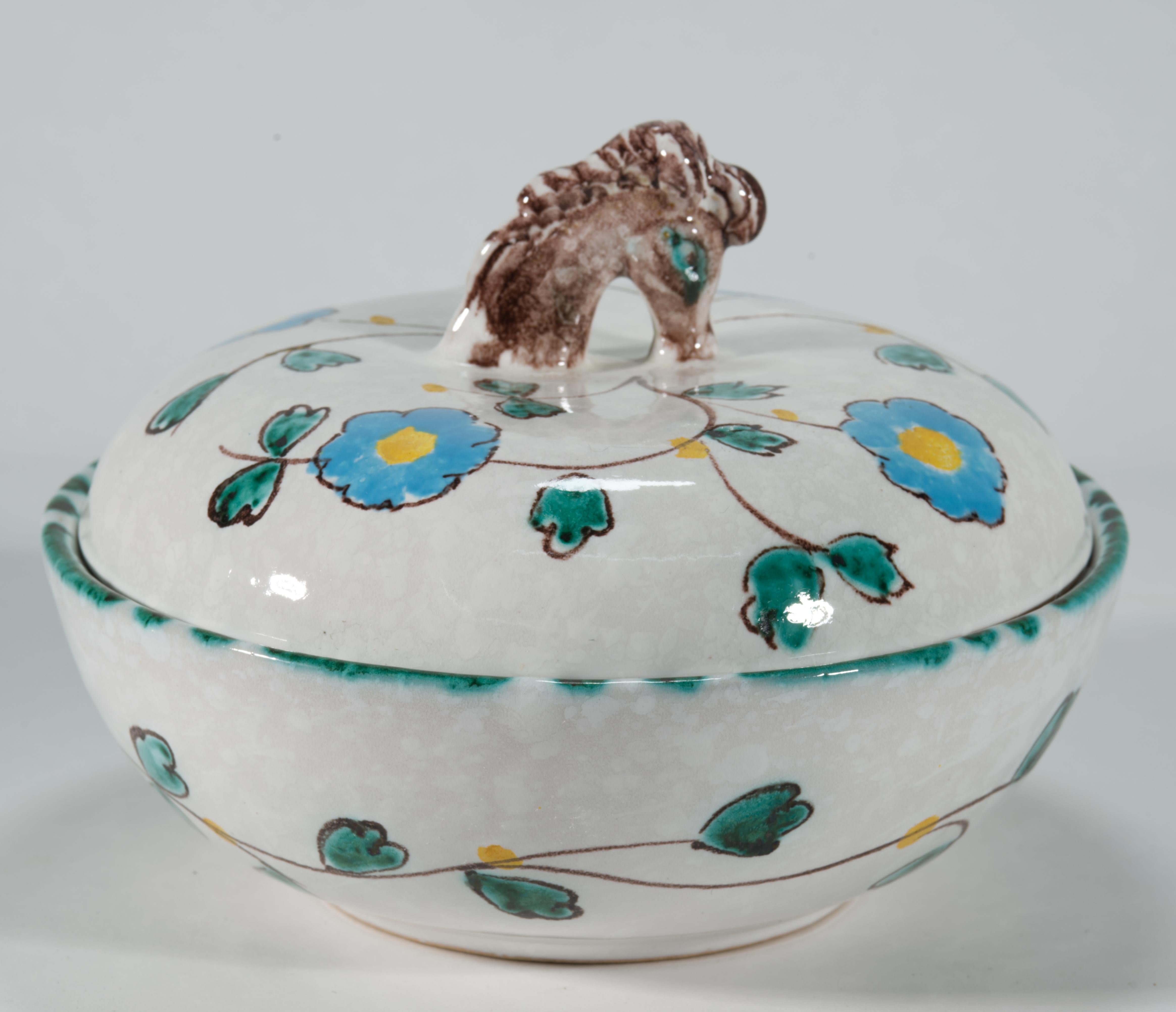  La cassettiera rotonda in ceramica maiolicata vintage con coperchio è decorata con fiori e foglie astratte dipinte a mano su sfondo bianco. I colori blu, verde acqua e marrone della decorazione floreale si ripetono nella decorazione del manico; il