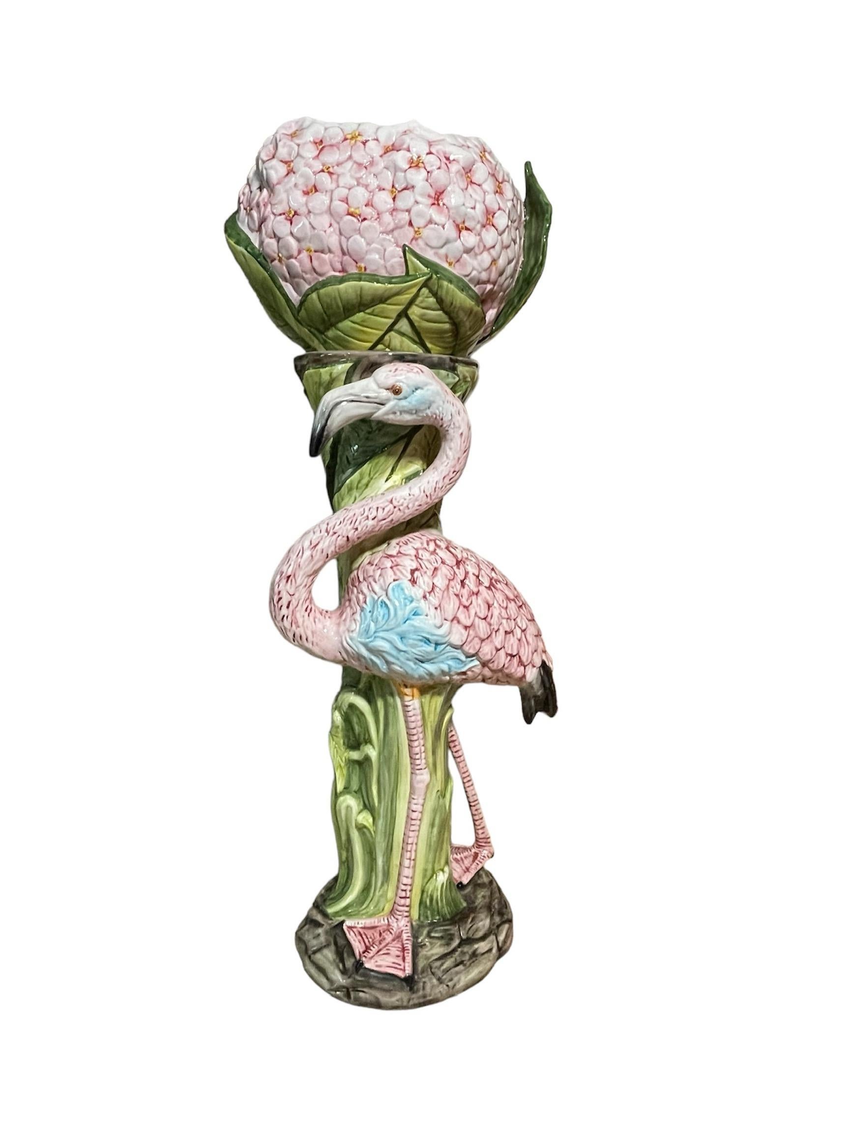 This is a Majolica Jardiniere and Pedestal. It depicts a Jardiniere decorated with a cluster of pink flowers that appears to be Hydrangeas at the bowl and its base adorned with large green leaves. The pedestal long “stem” is enfolded with the same