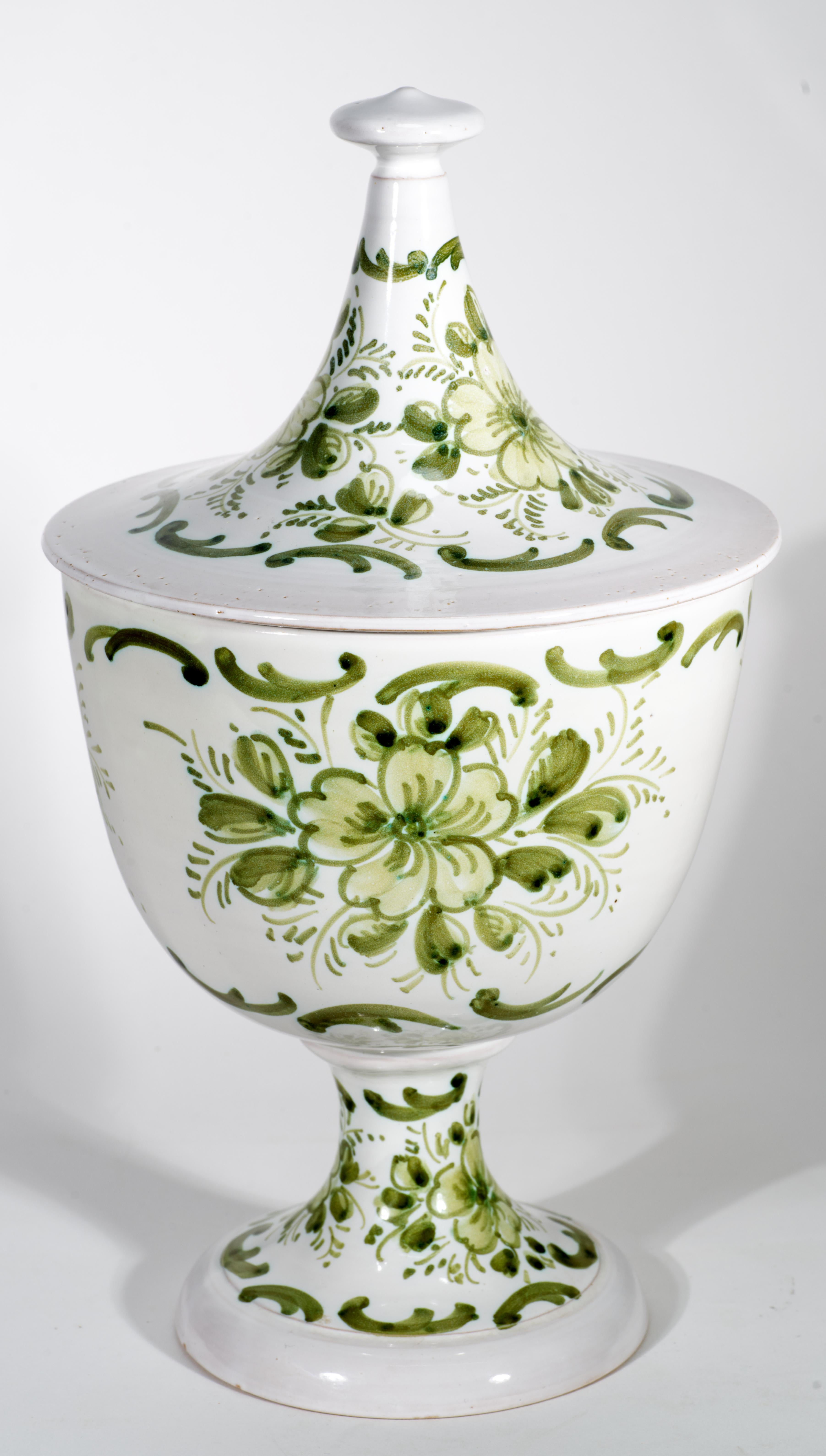 Vintage Majolica tall lidded footed urn vase features abstract floral and geometric decor in shades of green on white background. The vase stands on a rounded pedestal foot and is covered with circular lid accented with elongated handle adorned with