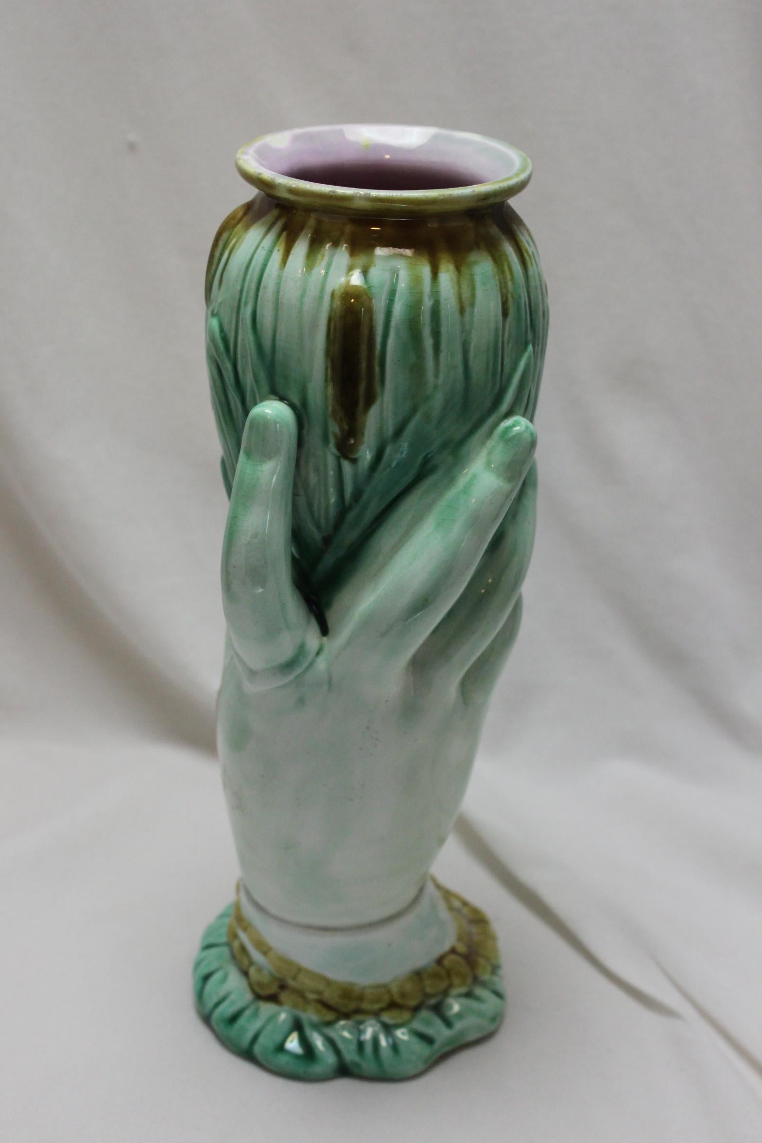 This majolica glazed vase is made up of a ladies hand holding a bunch of bullrushes which forms the vase part. Her wrist is clothed in a ruffled sleeve, over which is a beaded bracelet, and she has an unusual ring on her index finger. The vase is