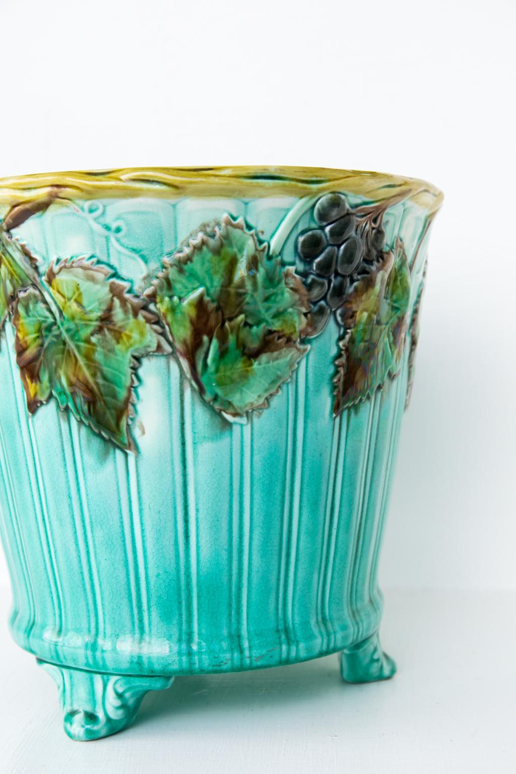 Majolica jardinière, has a ribbed body and molded acanthus feet. The top rim depicts a trailing grapevine with grapes and leaves. There is simulated basket weave in a yellow motif.
       