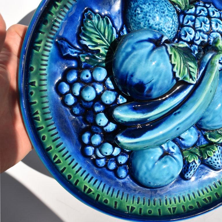 Round blue decorative Majolica fruit plate by Inarco. This decorative plate features a molded grouping of various fruits such as bananas, strawberries, grapes, apples, pineapples, peaches, oranges and berries. All glazed in a bright blue and green
