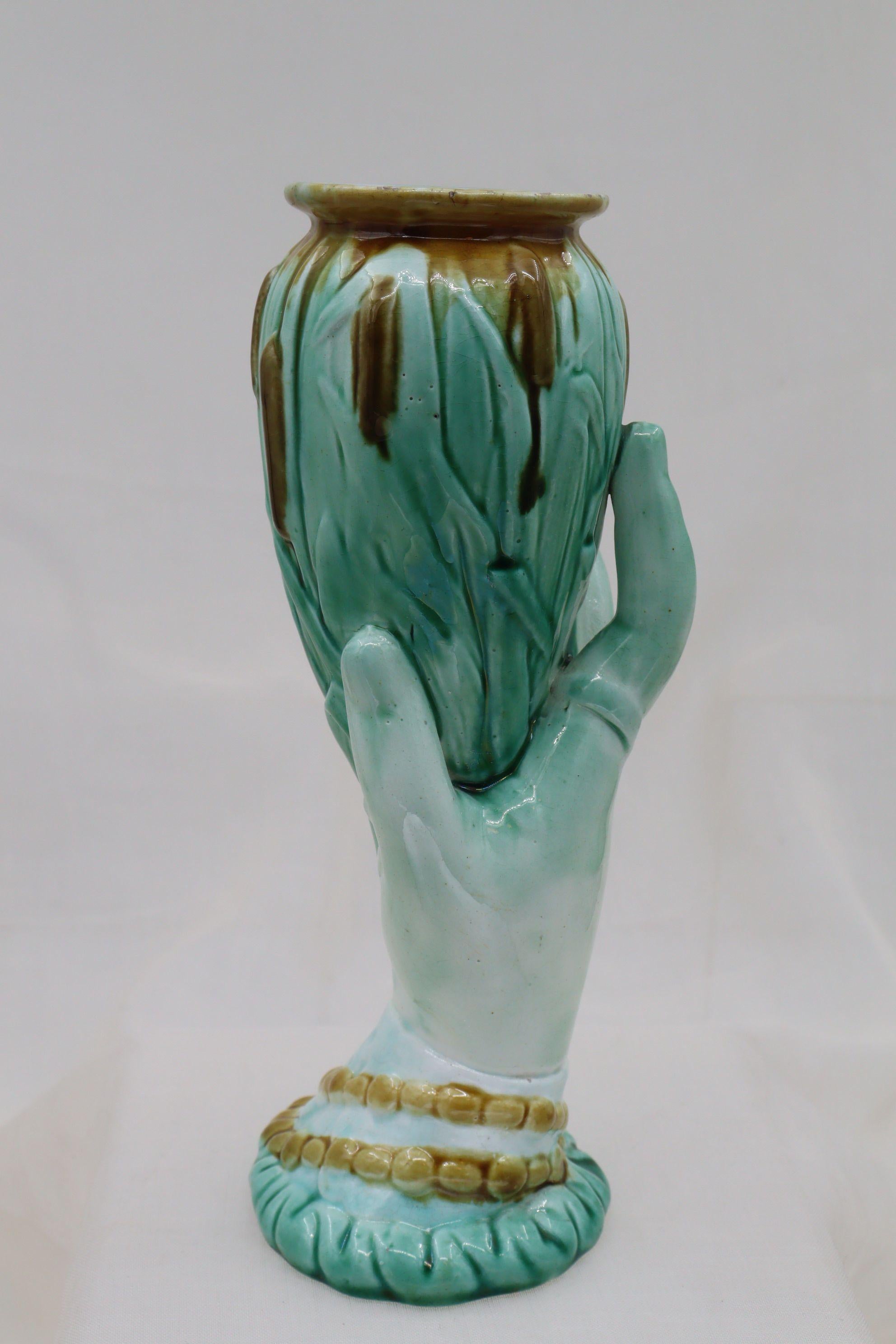 This majolica glazed vase is made up of a ladies hand holding aloft a bunch of bullrushes and cattails which forms the vase part. Her wrist is clothed in a ruffled sleeve, over which is a beaded bracelet, and she has an unusual ring on her index