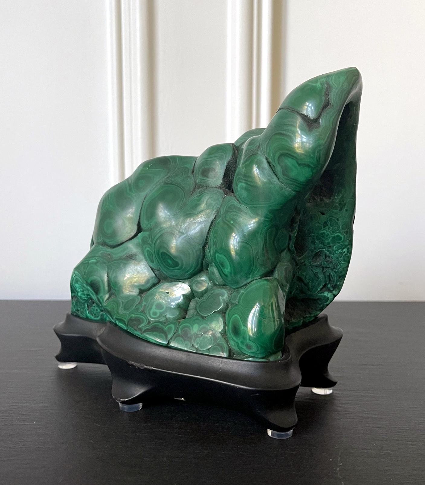A malachite rock specimen with intense green and black colors fitted on a wood stand. The gemstone in the botryoidal form was polished in all sides except one side with a fissure to reveal the beautiful swirling patterns and colors. It is displayed