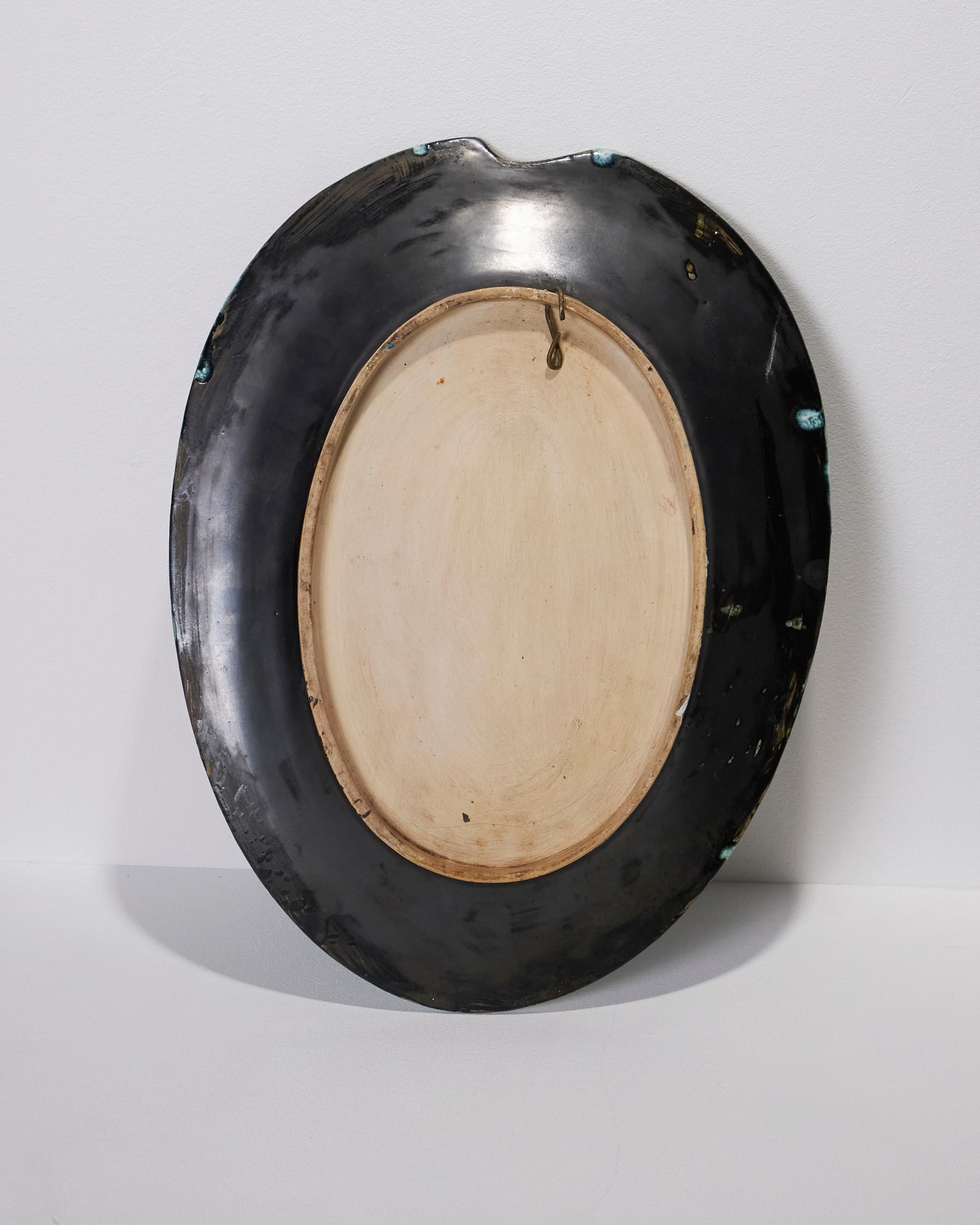 This decorative oval plate on a pedestal, a tribute to Malevich, features a small notch made before firing on the edge of the wing, directly in line with the face, to evoke a painter's palette. The design depicts a frontal face on a background of