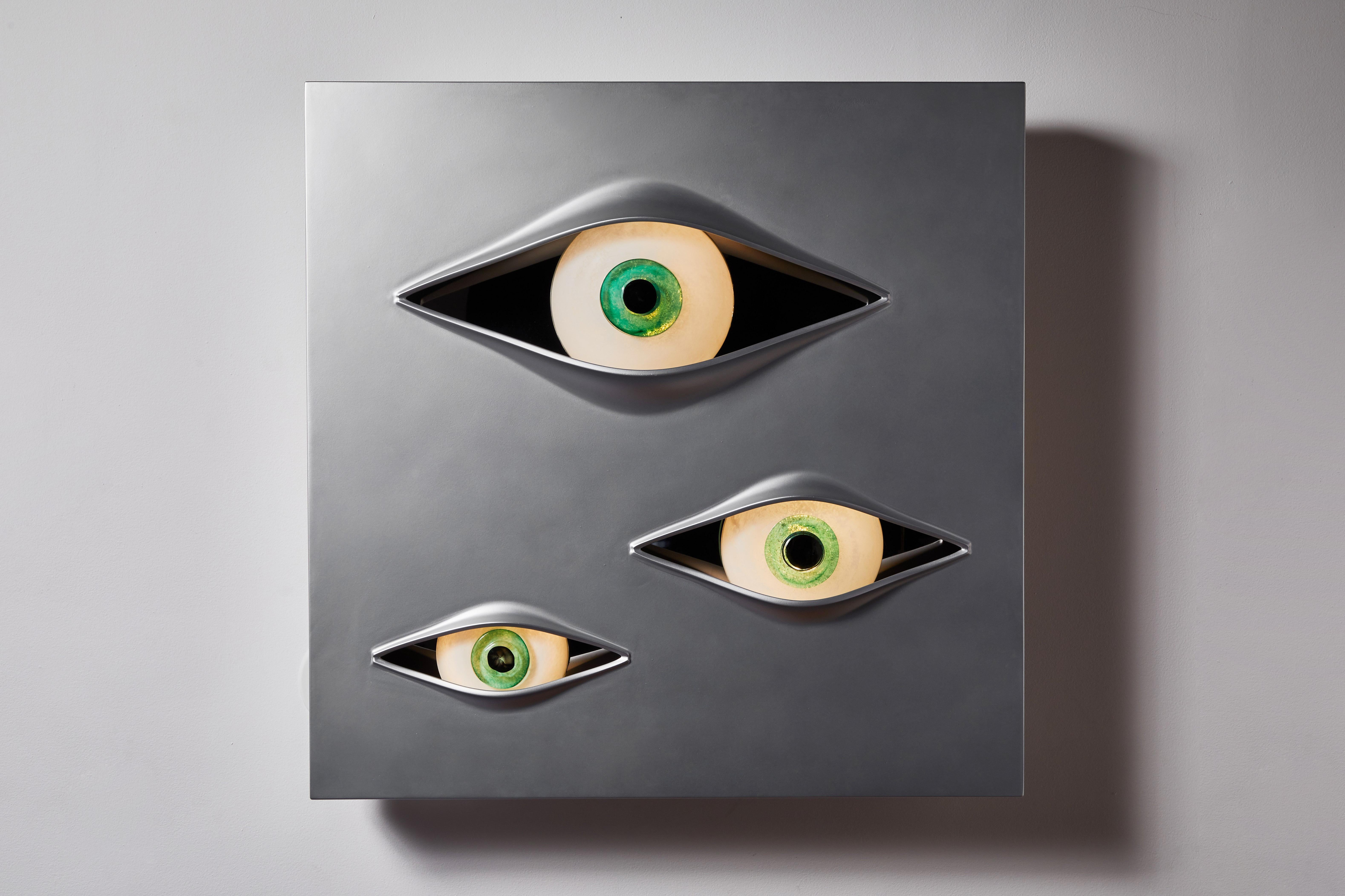 Malizia wall light by Angelo Brotto. Originally designed in 1973. Current production manufactured in Italy by Esperia. Luminous wall sculpture made with a panel of brushed aluminum and three eyes made of Murano glass. Takes three 11w LED bulbs.