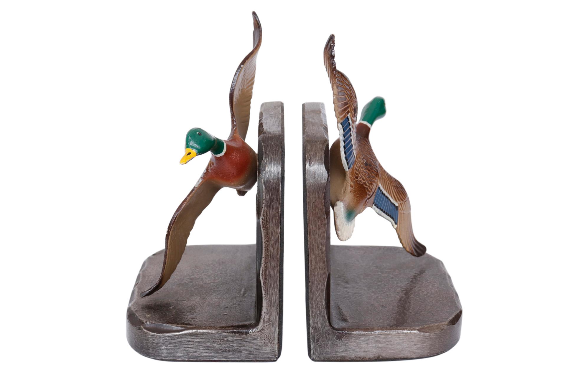A pair of metal bookends decorated with mallard ducks taking flight. Pressed with feather details and hand painted with the green and blue feathers particular to the mallard duck. Mounted on metal bases etched with a water scene. Dimensions per