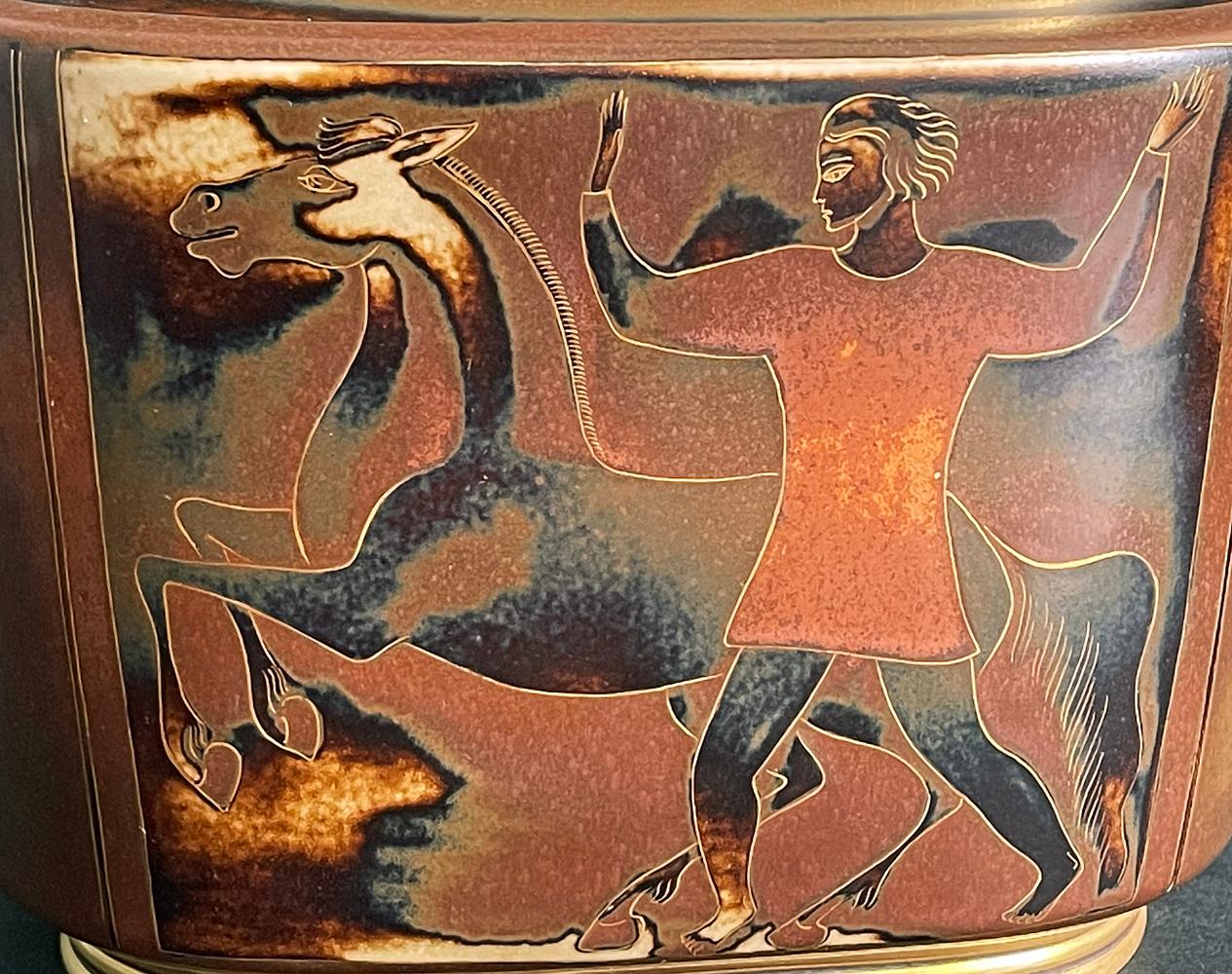 Gorgeously glazed in tones of rust, sable, ivory and chocolate, this unique lidded urn with foliated pinnacle and curving handles depicts a man with upraised arms and rearing horse on one side, and a female figure riding a horse on the other. The