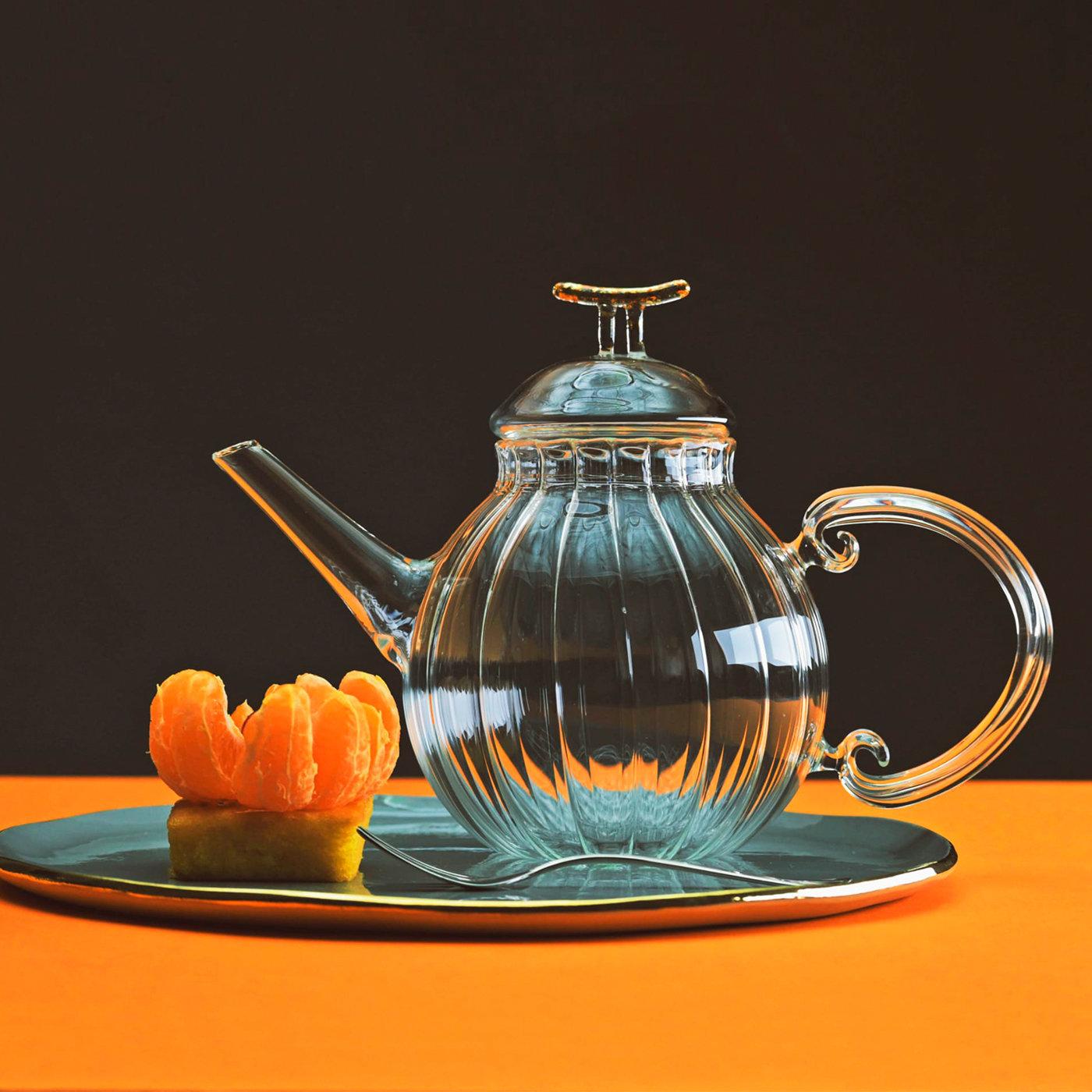 Diese elegante Teekanne wird vollständig von Hand aus transparentem, mundgeblasenem Glas hergestellt. Sie besticht durch eine auffällige Textur aus parallelen Linien, die vertikal auf dem geschwungenen Körper verlaufen und die geschwungene Form