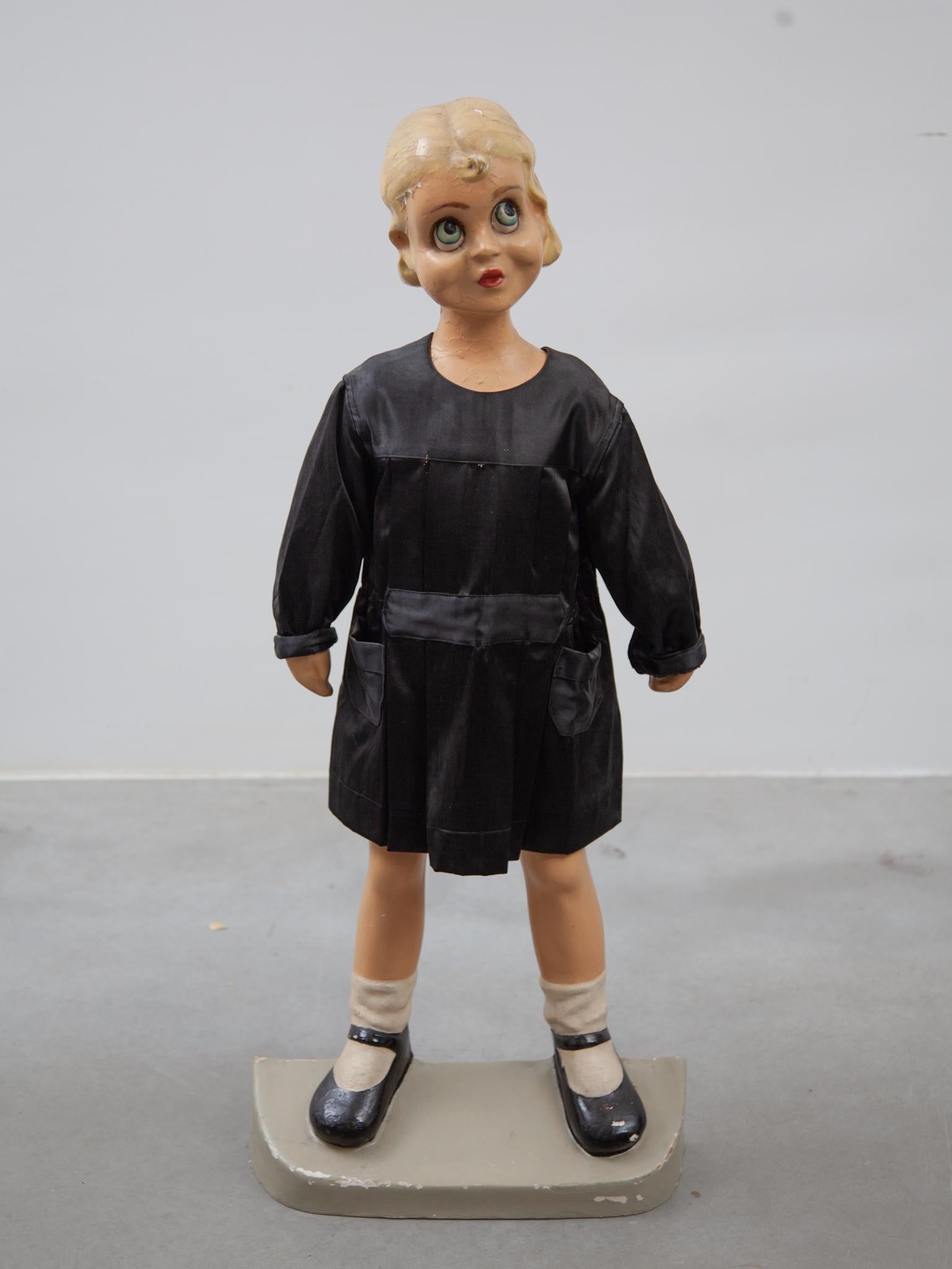 Original Schaufensterpuppe für Grundschulkleidung, sie trägt ein schwarzes Kleid als Beispiel für Kinderuniformen aus den 1950er Jahren.