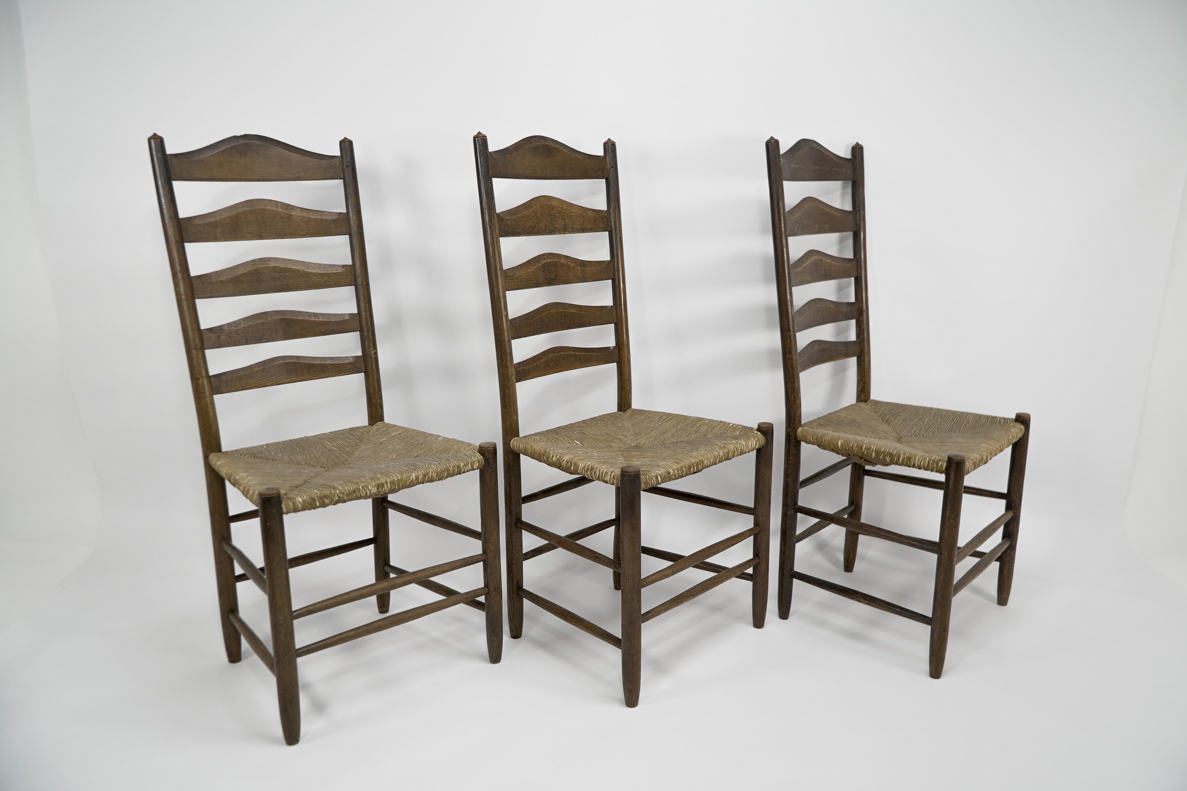 À la manière d'E Gimson. Un ensemble de trois chaises de salle à manger ou d'appoint en chêne de qualité Arts and Crafts avec des échelles sculptées à la main à l'arrière et conservant les sièges en jonc d'origine. Prix de l'ensemble
