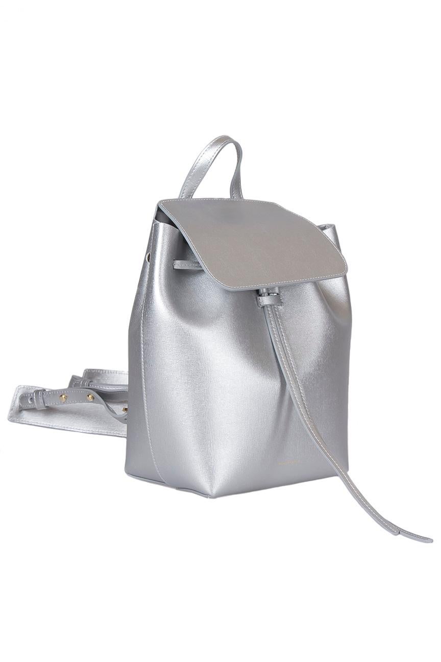 This stylish backpack from Mansur Gavriel is crafted from silver leather and enhanced with gold-tone hardware. The bag features a top handle and adjustable shoulder straps. A flap secures the drawstring closure that opens to reveal a spacious
