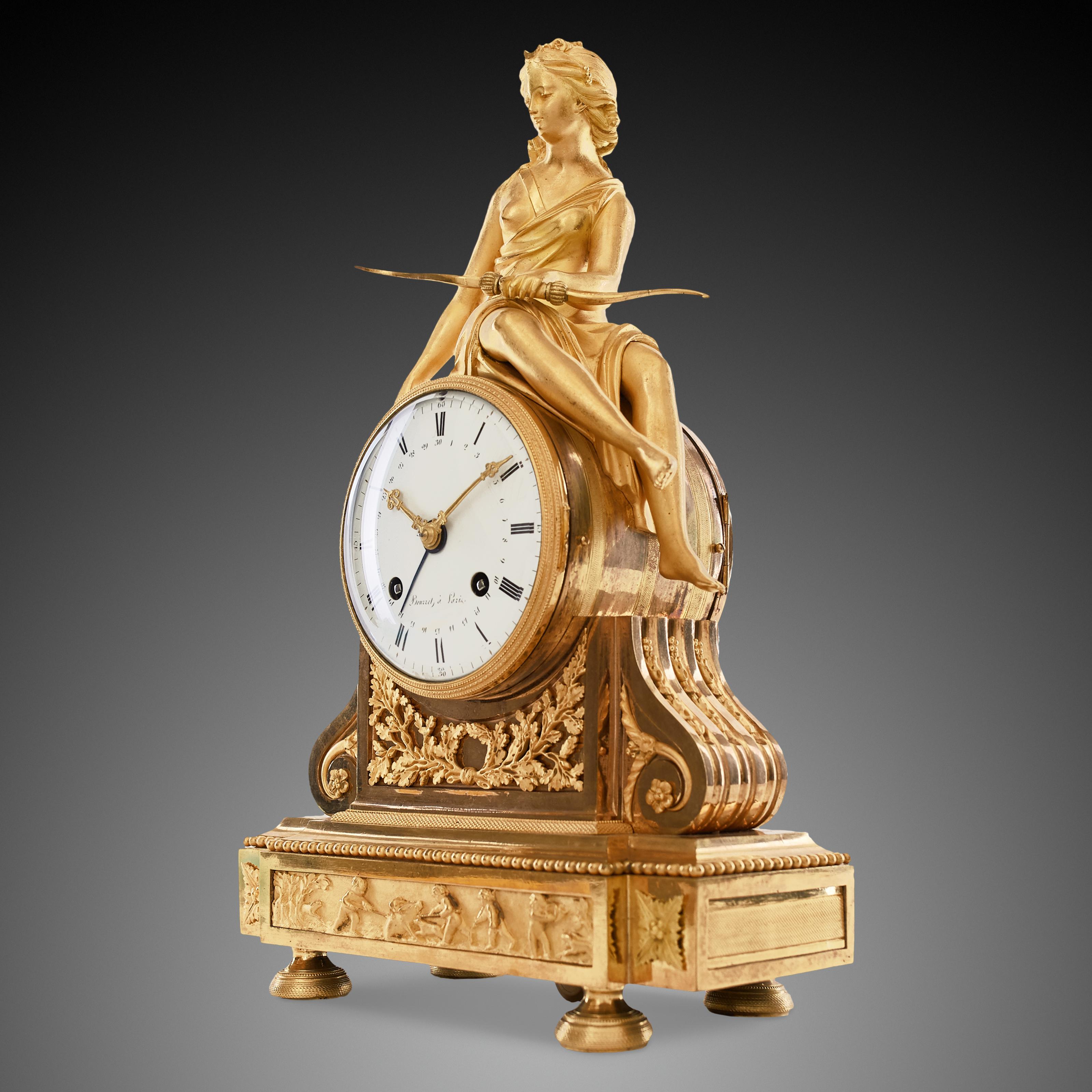 The mantel clock, made of ormol (gilded bronze), shows the beautiful figure of Diana, the Roman goddess of the hunt, accompanied by various emblems. Diana is sitting on the circular clock envelope, holding an arrow with her left hand and a dead bird