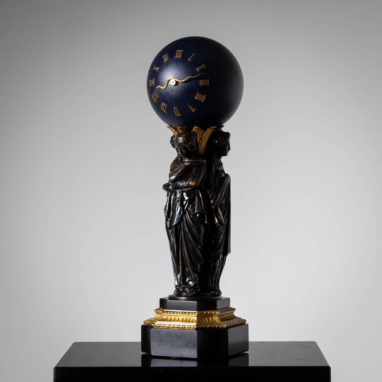 Bronze clock with a dark blue, spherical clock case with Roman hours and snake-shaped hands. The case is supported by three female personifications of the arts of painting, sculpture and architecture. They stand on a curved stone base with a gilded