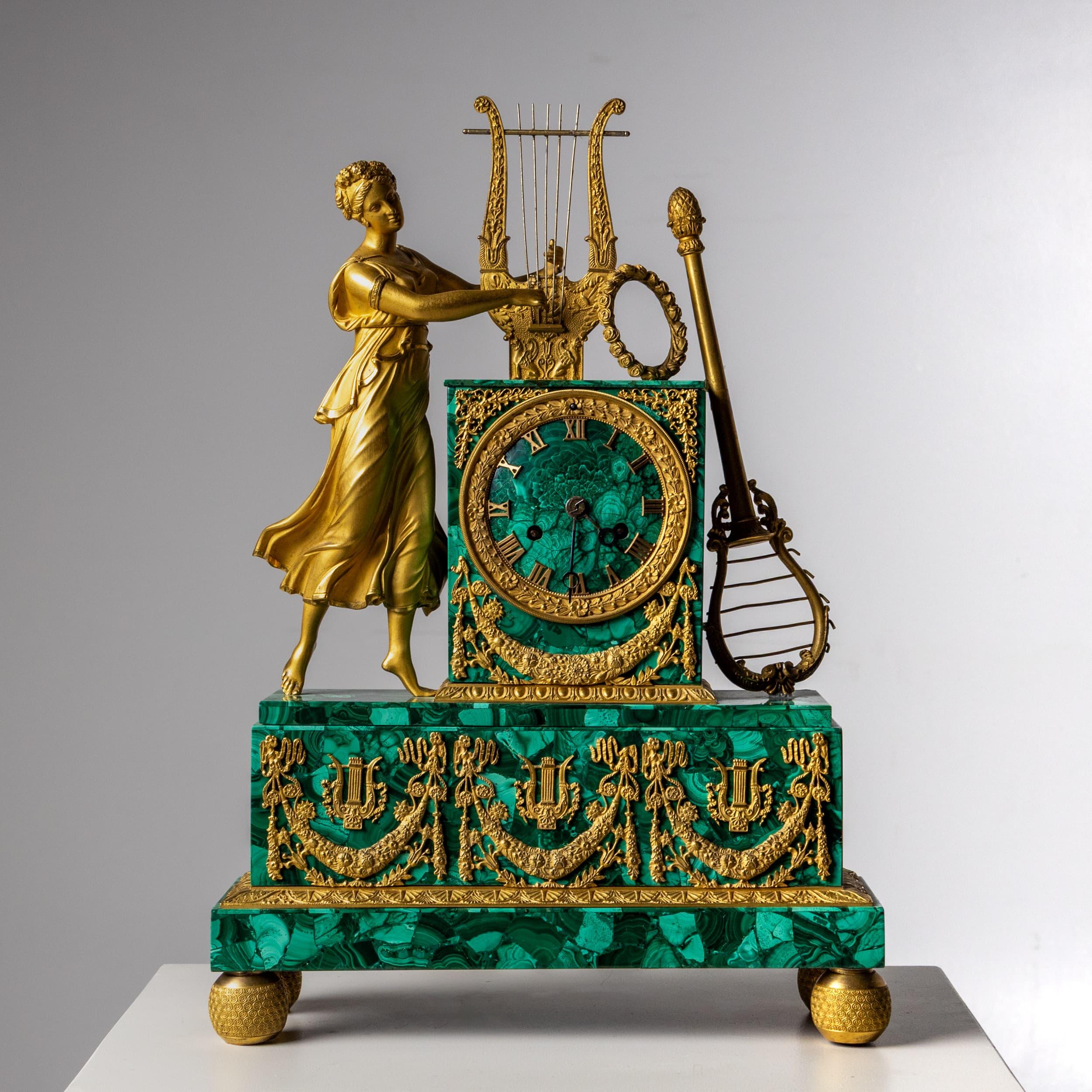 This elegant mantel clock features a rectangular case entirely clad in deep green malachite. Fire-gilded bronze decorations in the form of floral festoons and lyres adorn the case. The clock is crowned by a fully sculpted figure of a young woman